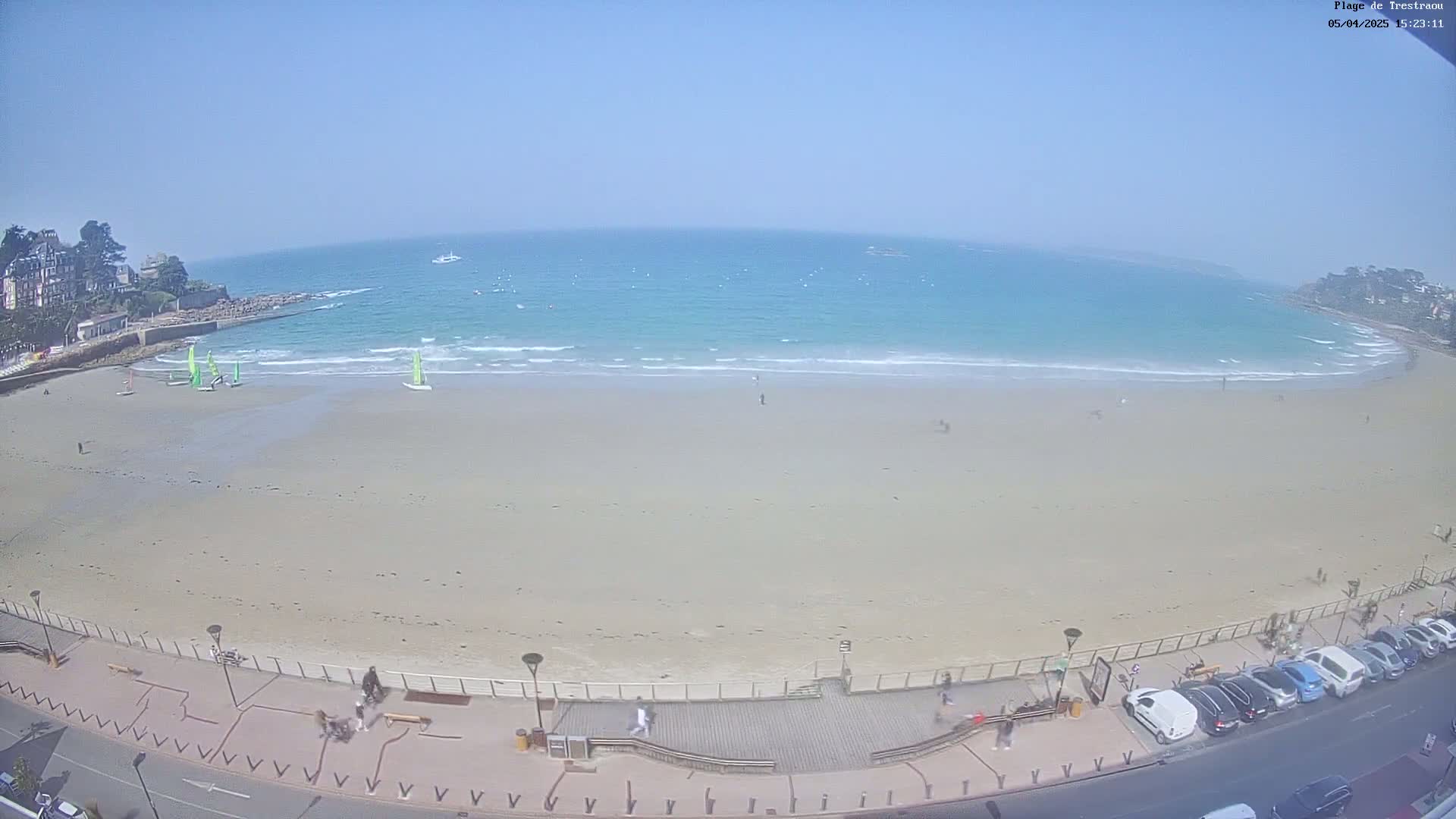 A sandy beach with a calm, turquoise ocean under a clear blue sky is viewed from above, showing several people and sailboats near the shore, and cars parked along the road adjacent to the beach.