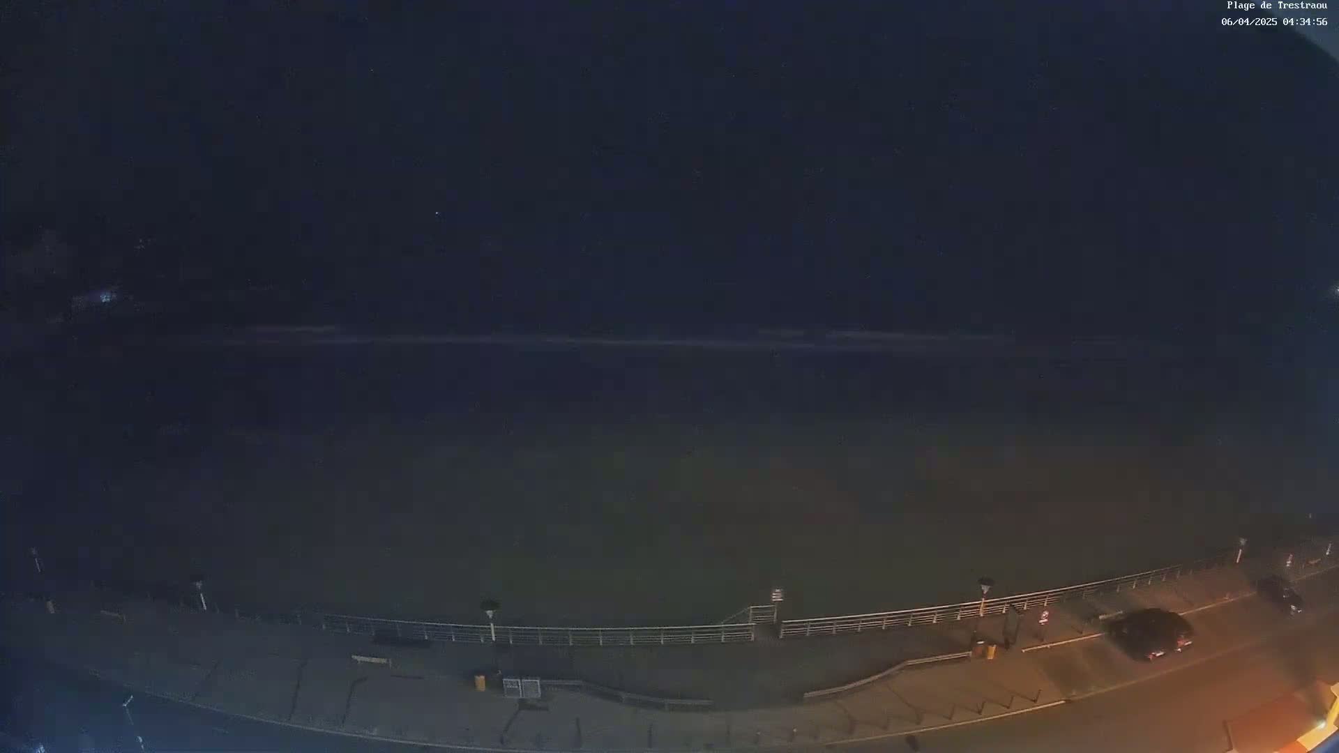 A nighttime, high-angle view shows a dark body of water next to a paved walkway with a railing and a few parked cars under a clear, dark sky.