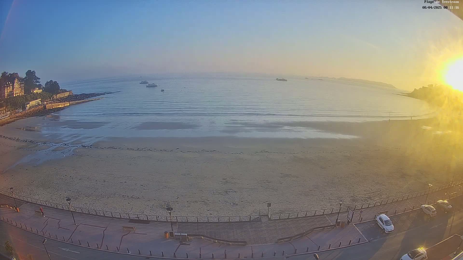 A sandy beach with calm waves and several boats in the distance, under a clear, sunny sky.