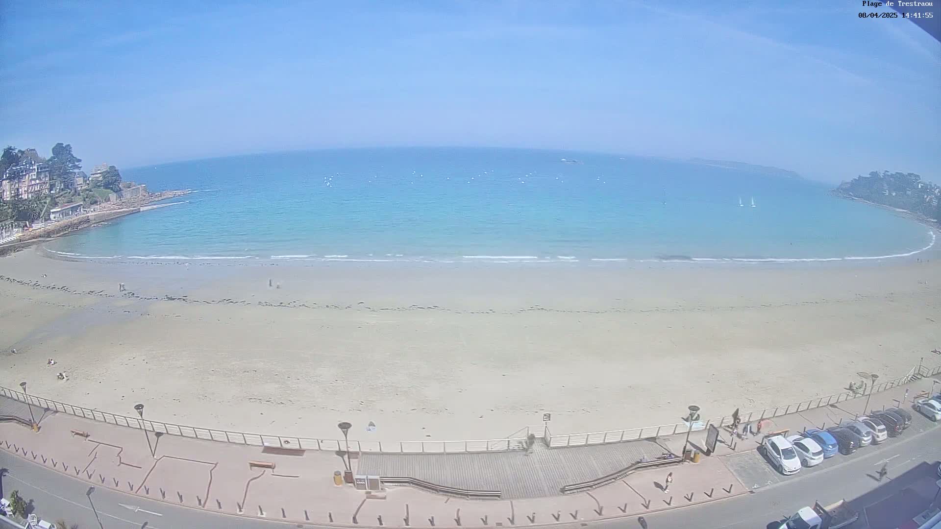 A sandy beach curves around a calm, light-blue ocean under a clear, sunny sky.