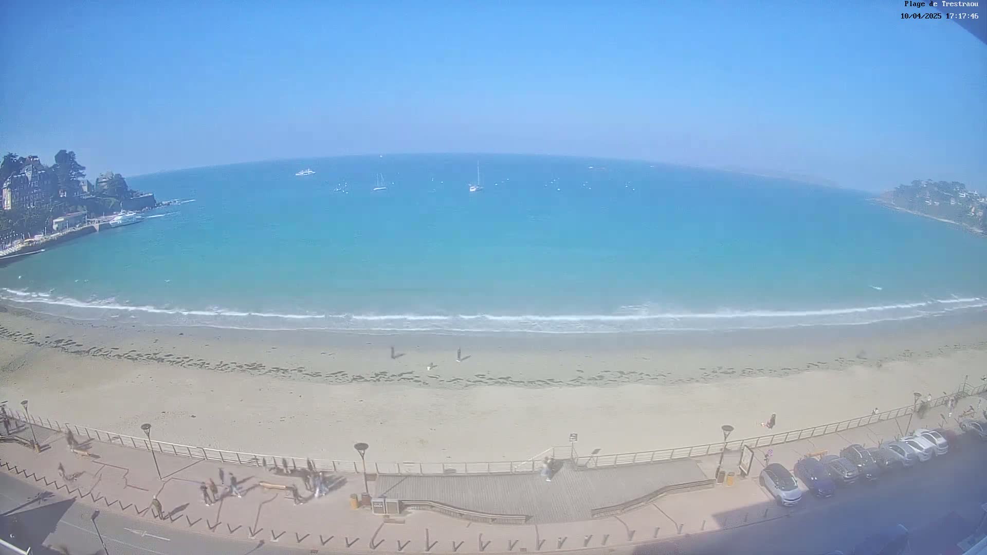 A sandy beach with a boardwalk and parked cars is visible, overlooking a calm, turquoise ocean under a clear blue sky.