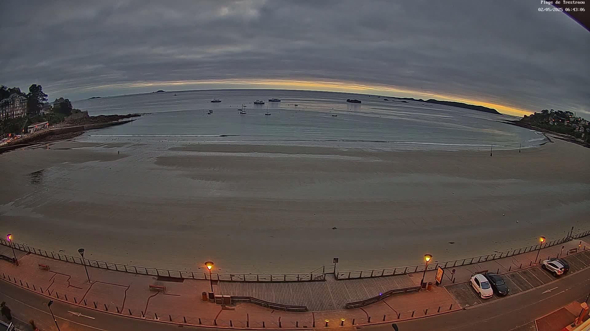 A wide shot of a sandy beach at dawn under an overcast sky, with several boats visible in the calm water and a few cars parked along a paved road adjacent to the beach.
