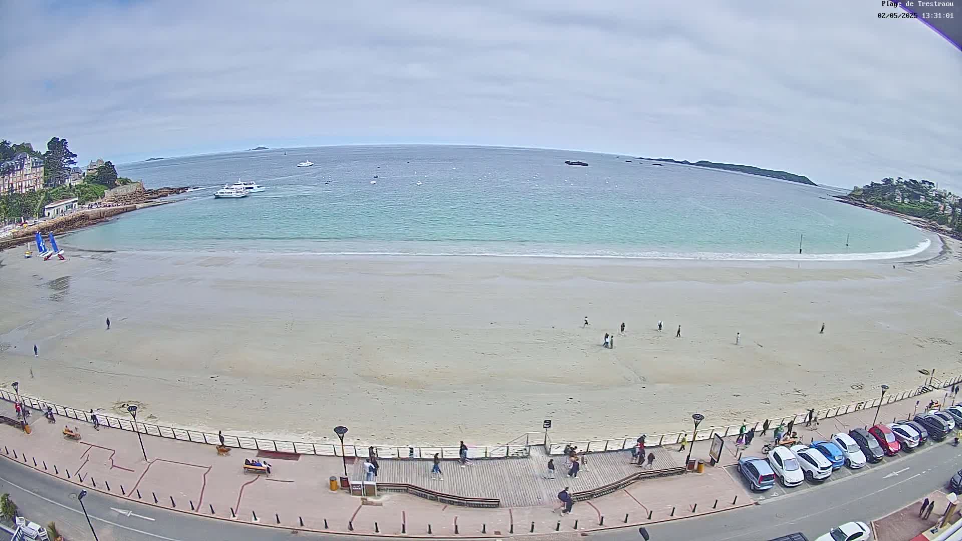 A sandy beach with light-colored sand and calm, light teal water is bordered by a paved walkway with people strolling and cars parked along the roadside under an overcast sky.