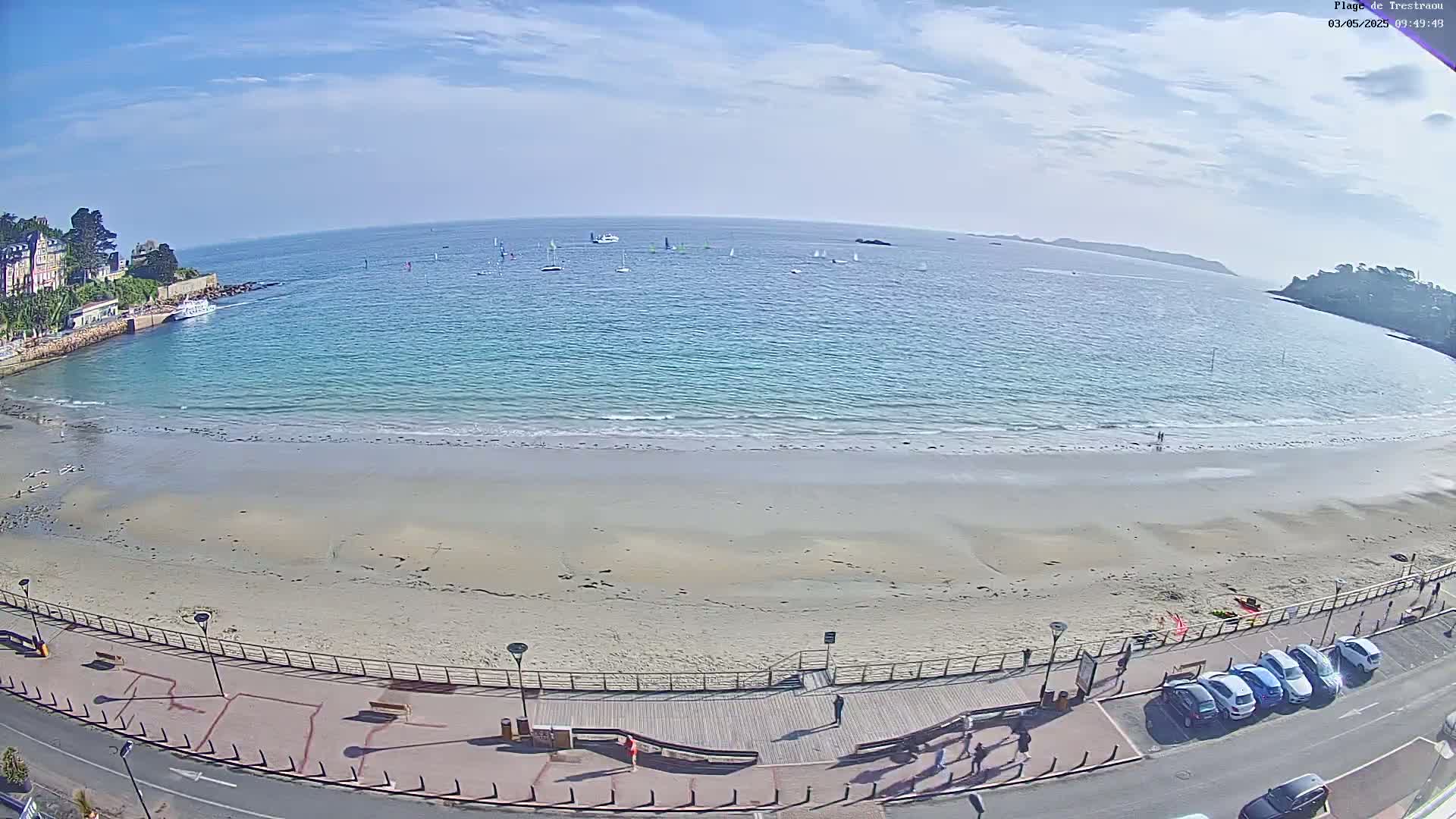A sandy beach with calm, light-blue water and several sailboats in the distance under a mostly clear sky is viewed from above.