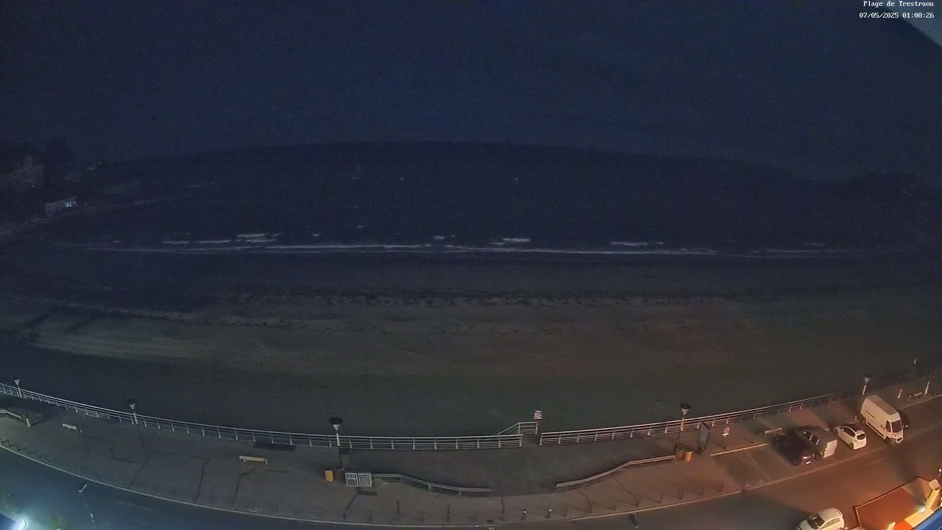A dark beach at night with gently lapping waves and a few vehicles parked along a nearby road.