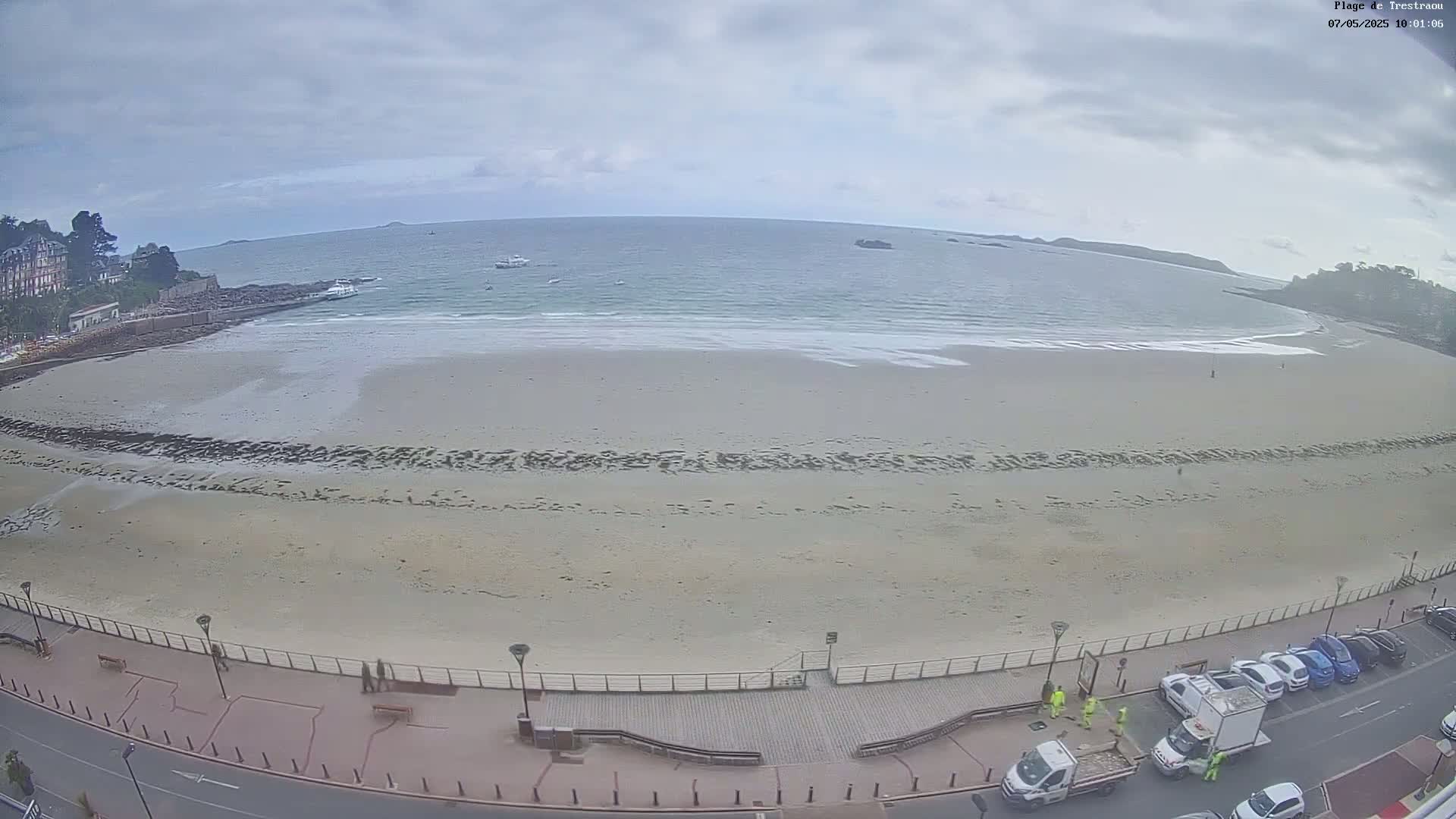 A mostly empty sandy beach stretches along a calm ocean under an overcast sky, with a boardwalk, parked cars, and a few people visible near the road.