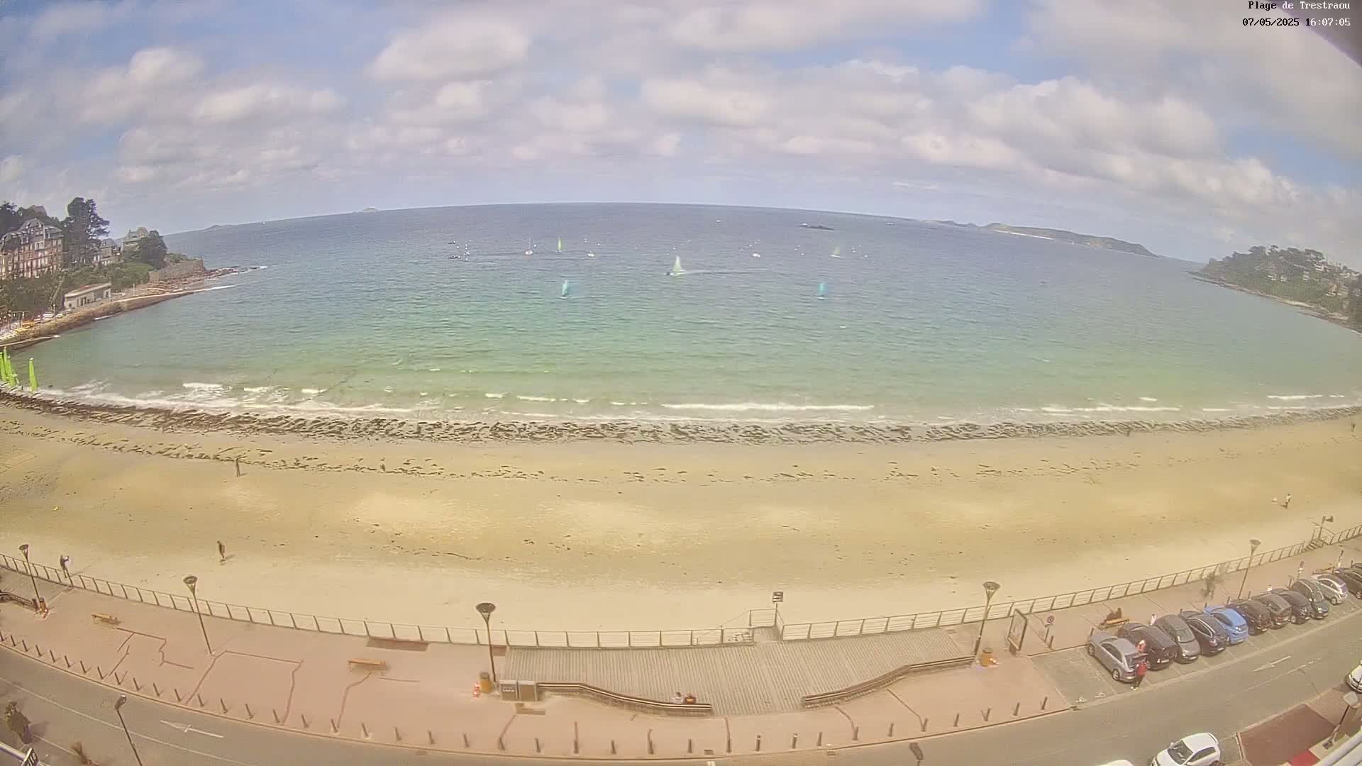 A sandy beach with several sailboats in the distance on a partly cloudy day, and cars parked along a road next to a boardwalk.