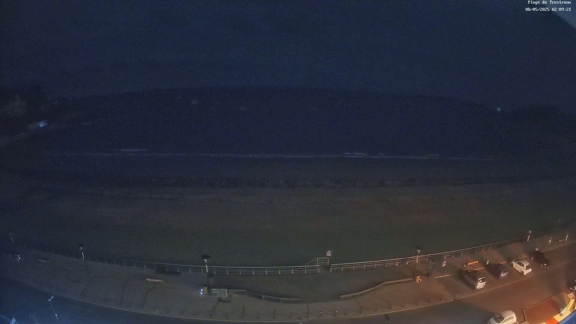 A dark, nighttime view shows a beach with calm waves and a few cars parked along a paved area next to a railing.