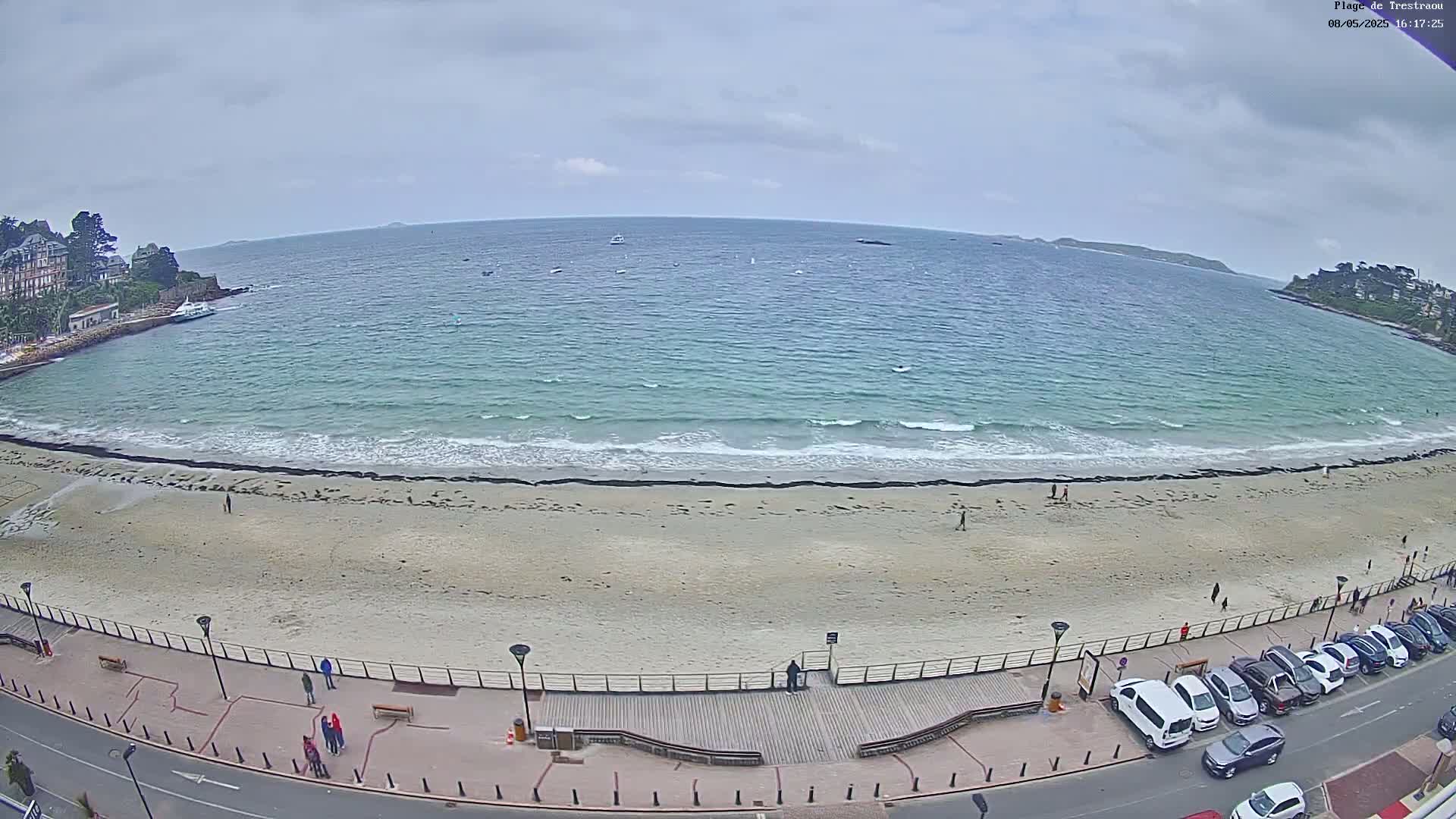 A sandy beach with gentle waves under a cloudy sky is bordered by a paved walkway lined with parked cars and buildings.