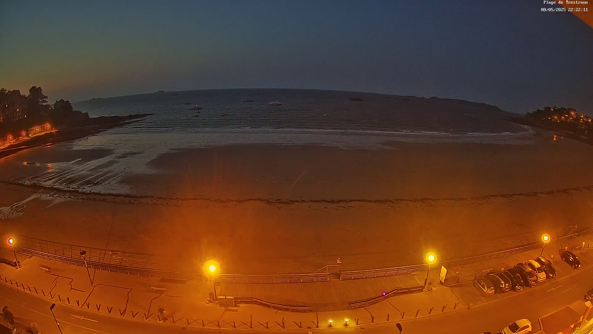 A wide, sandy beach at twilight is illuminated by streetlights along a boardwalk, with a calm ocean and several small boats visible in the distance.