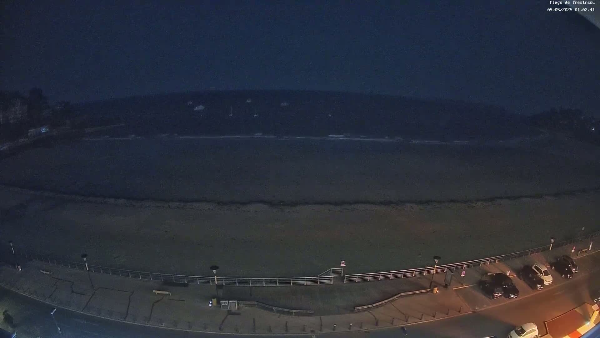 A nighttime, high-angle view shows a dark beach, calm water with a few boats visible in the distance, a boardwalk with parked cars, and streetlights illuminating the scene.