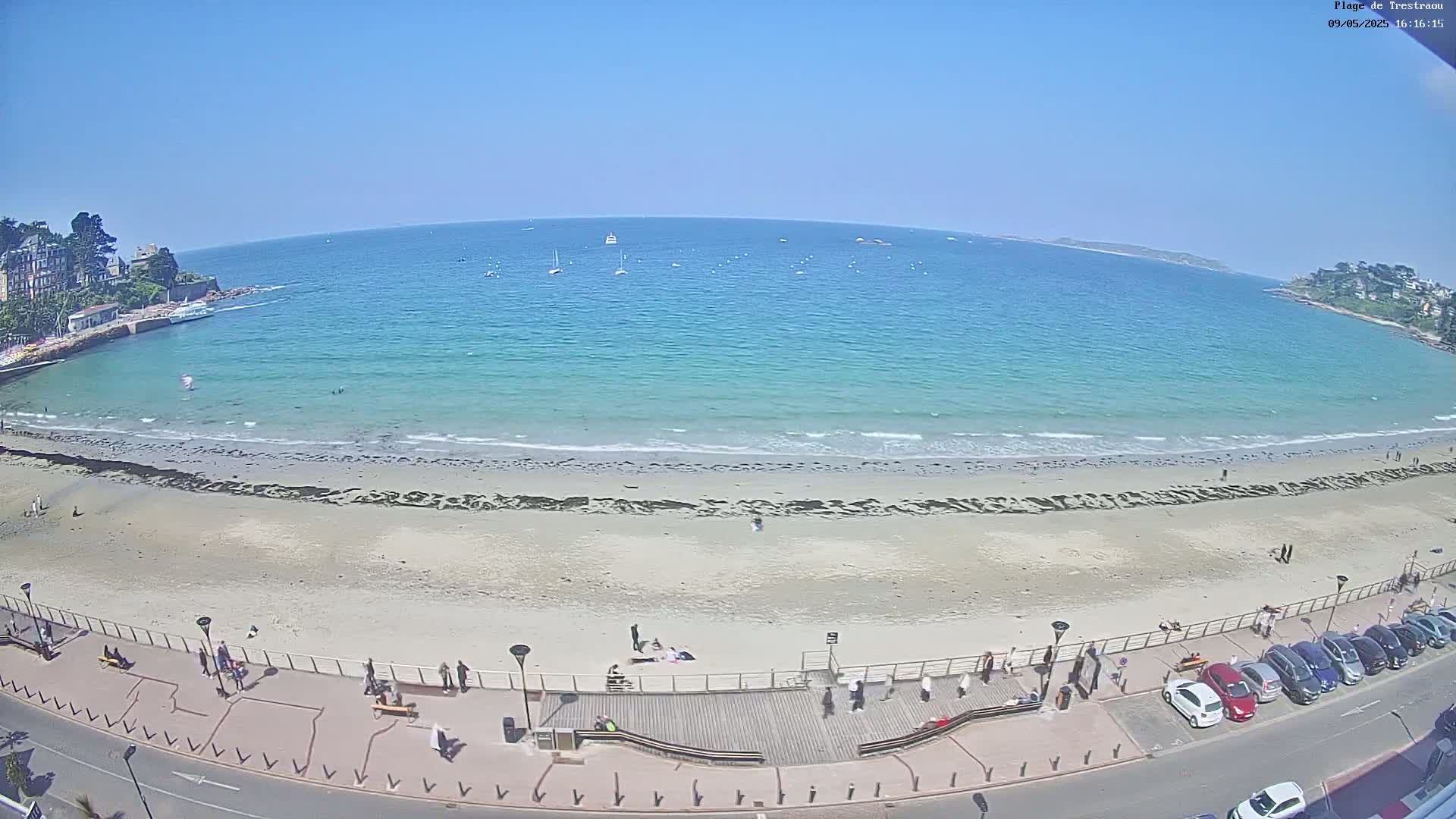 A sandy beach with several people on it and a calm, turquoise ocean under a clear blue sky is viewed from above.