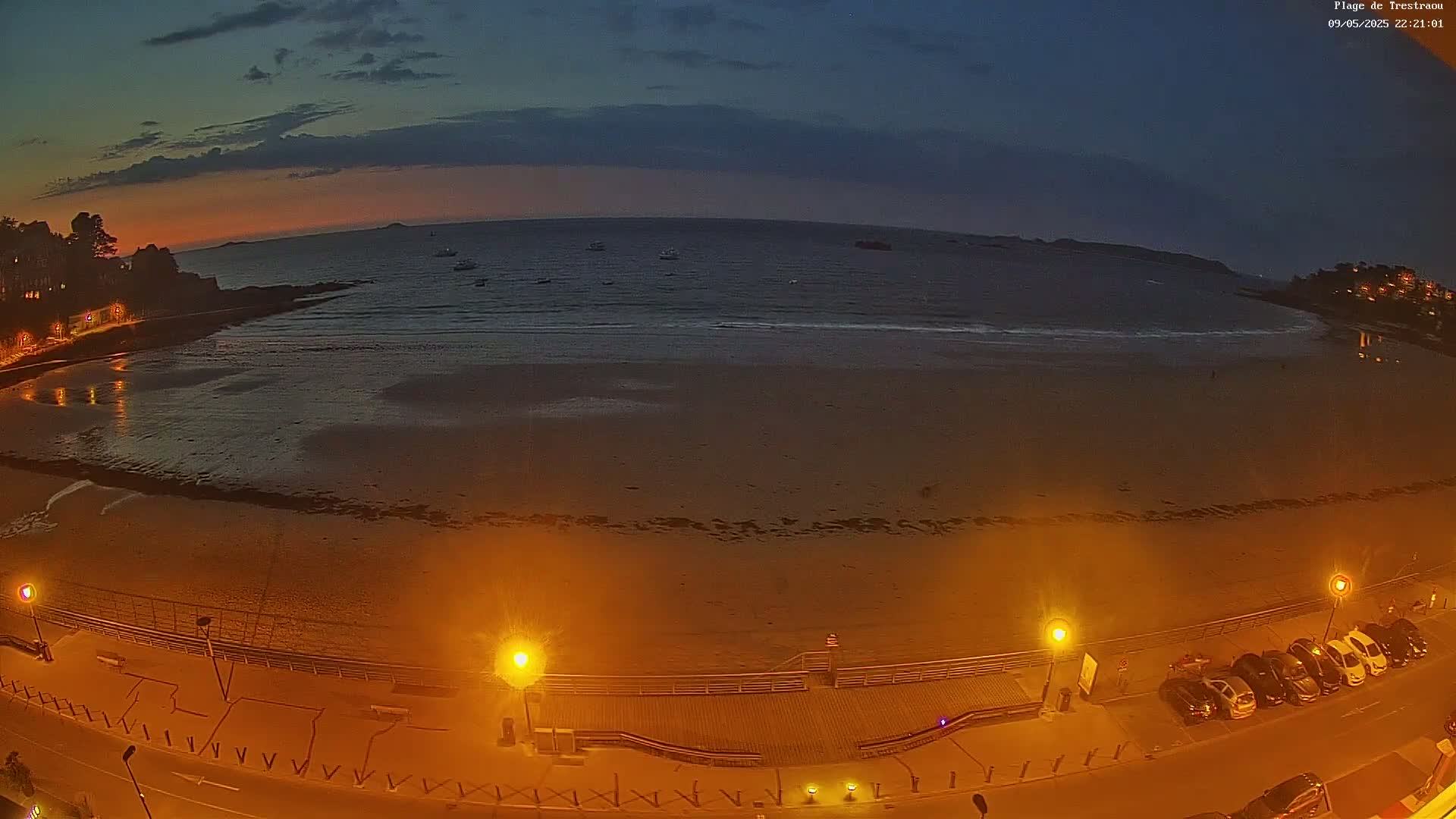 A mostly dark sandy beach at twilight is illuminated by streetlights, with a calm sea and a few boats visible in the distance.