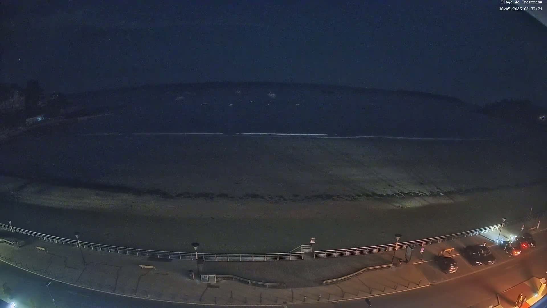 A dark, nighttime view shows a beach with a calm ocean, a boardwalk, and several cars parked along a road.