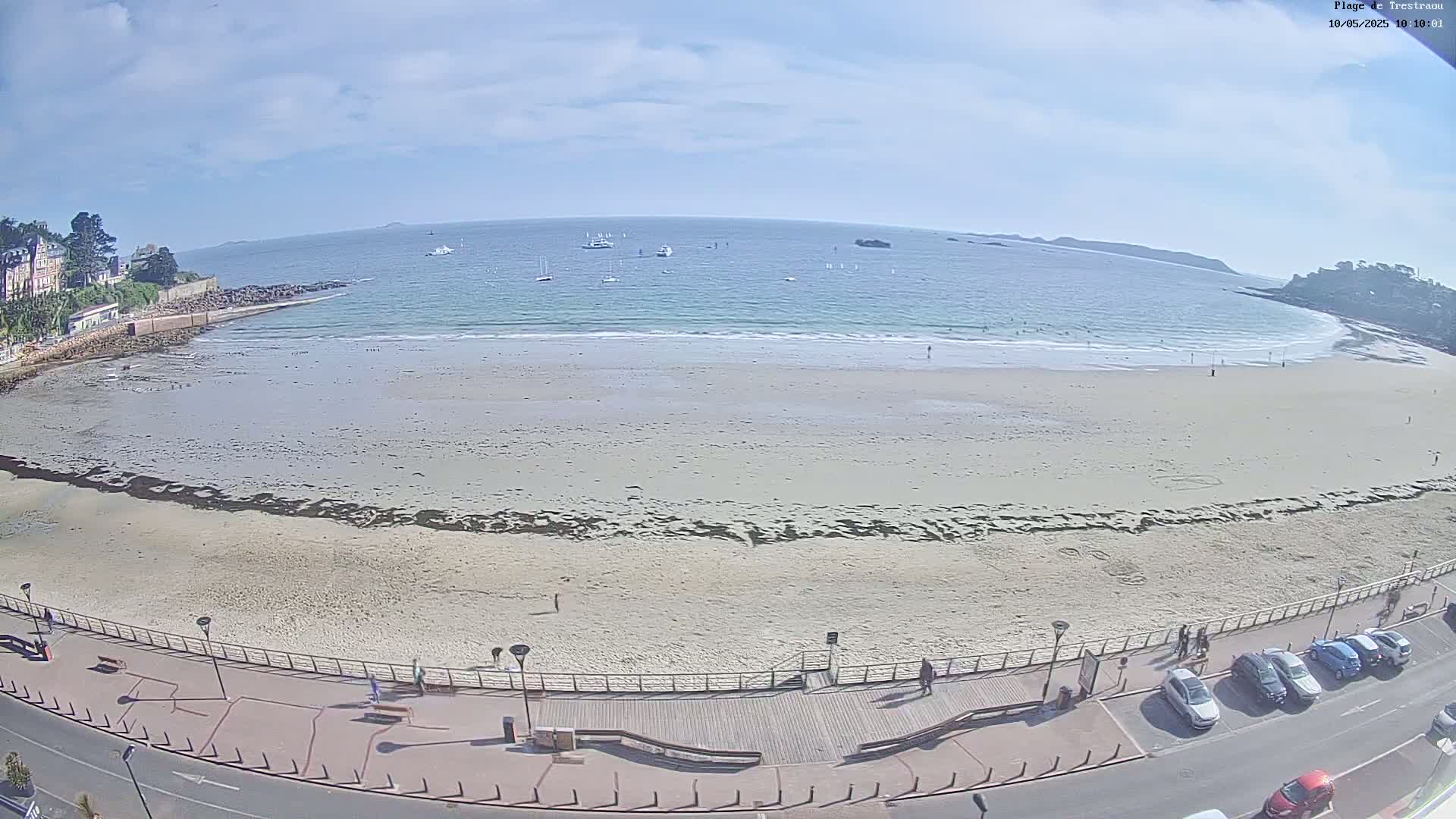 A sandy beach with a few people and several boats in the calm ocean under a partly cloudy sky is seen from an elevated vantage point.