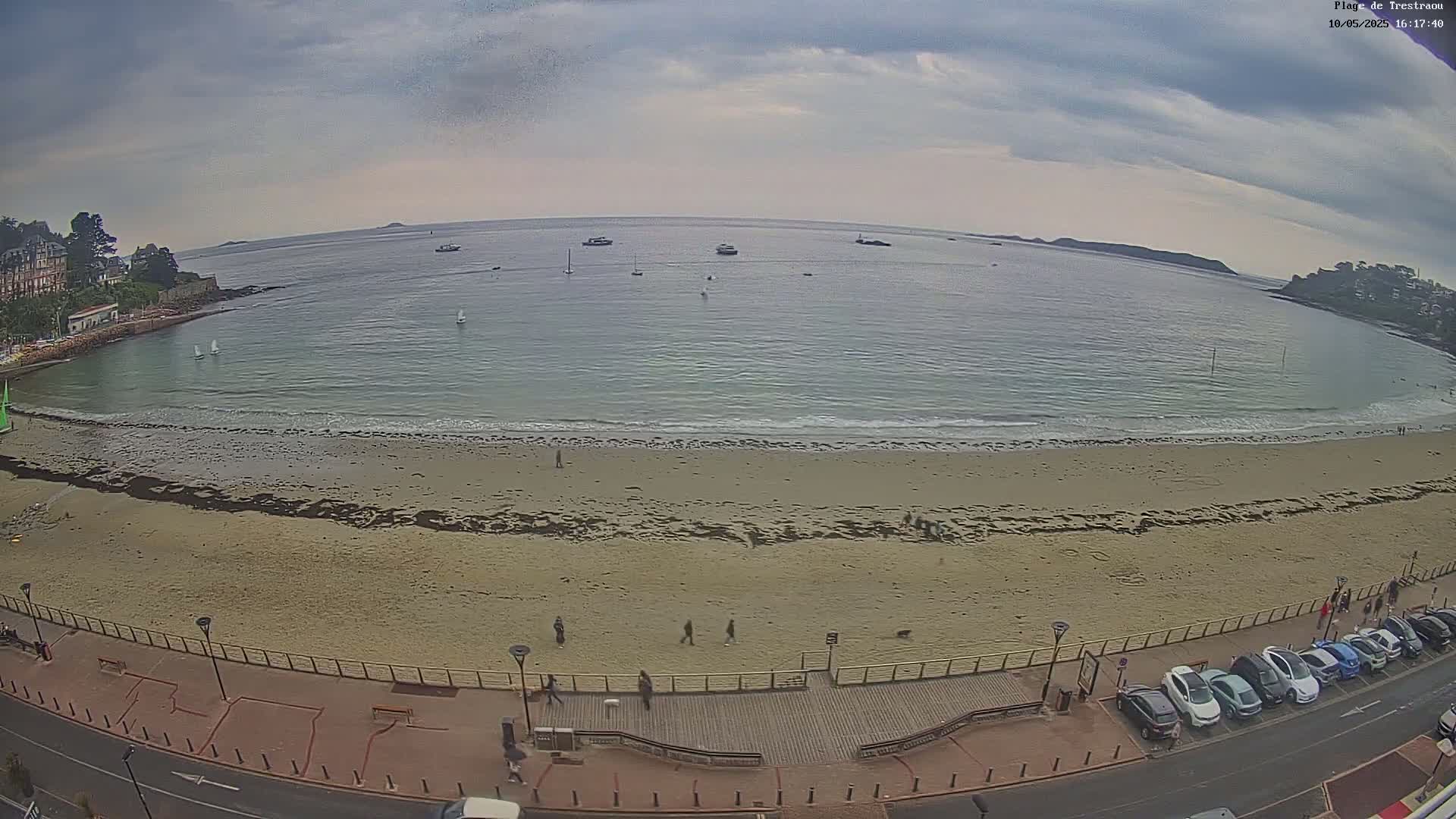 A sandy beach with several people walking along the shoreline and cars parked nearby, overlooked by buildings, under a mostly cloudy sky with small waves breaking on the shore and several sailboats visible in the calm bay.