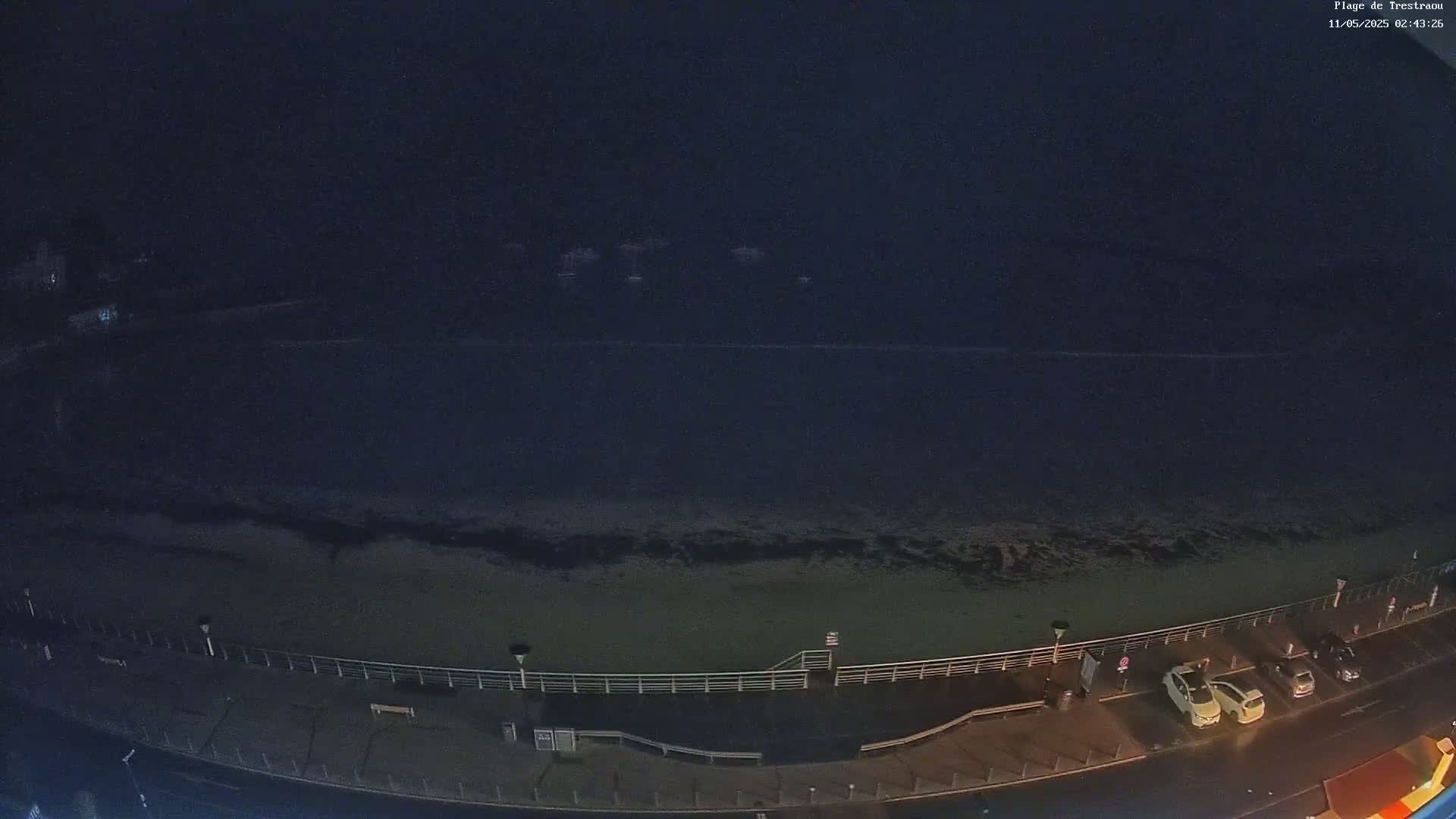 A dark, nighttime view of a beach with a few cars parked along a boardwalk, overlooking a calm, dark ocean.