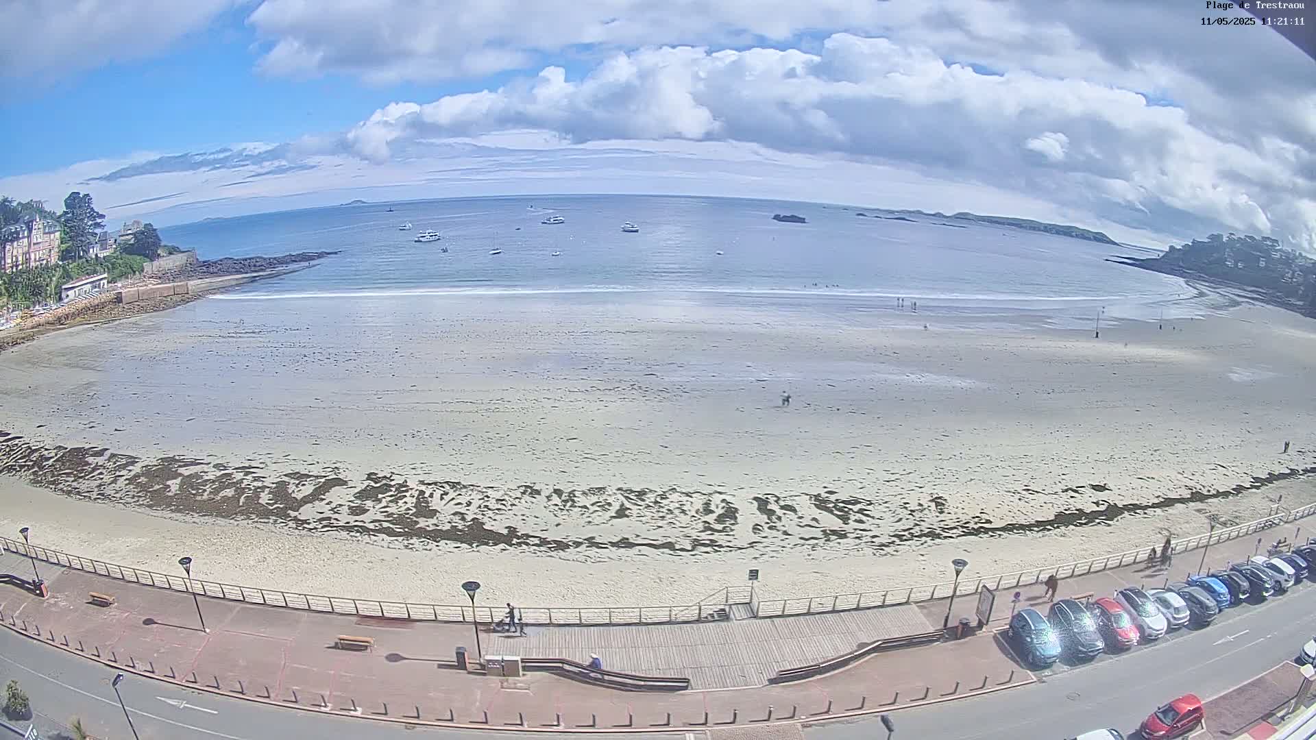 A wide sandy beach with a few people and some boats in the distance is visible under a partly cloudy sky.