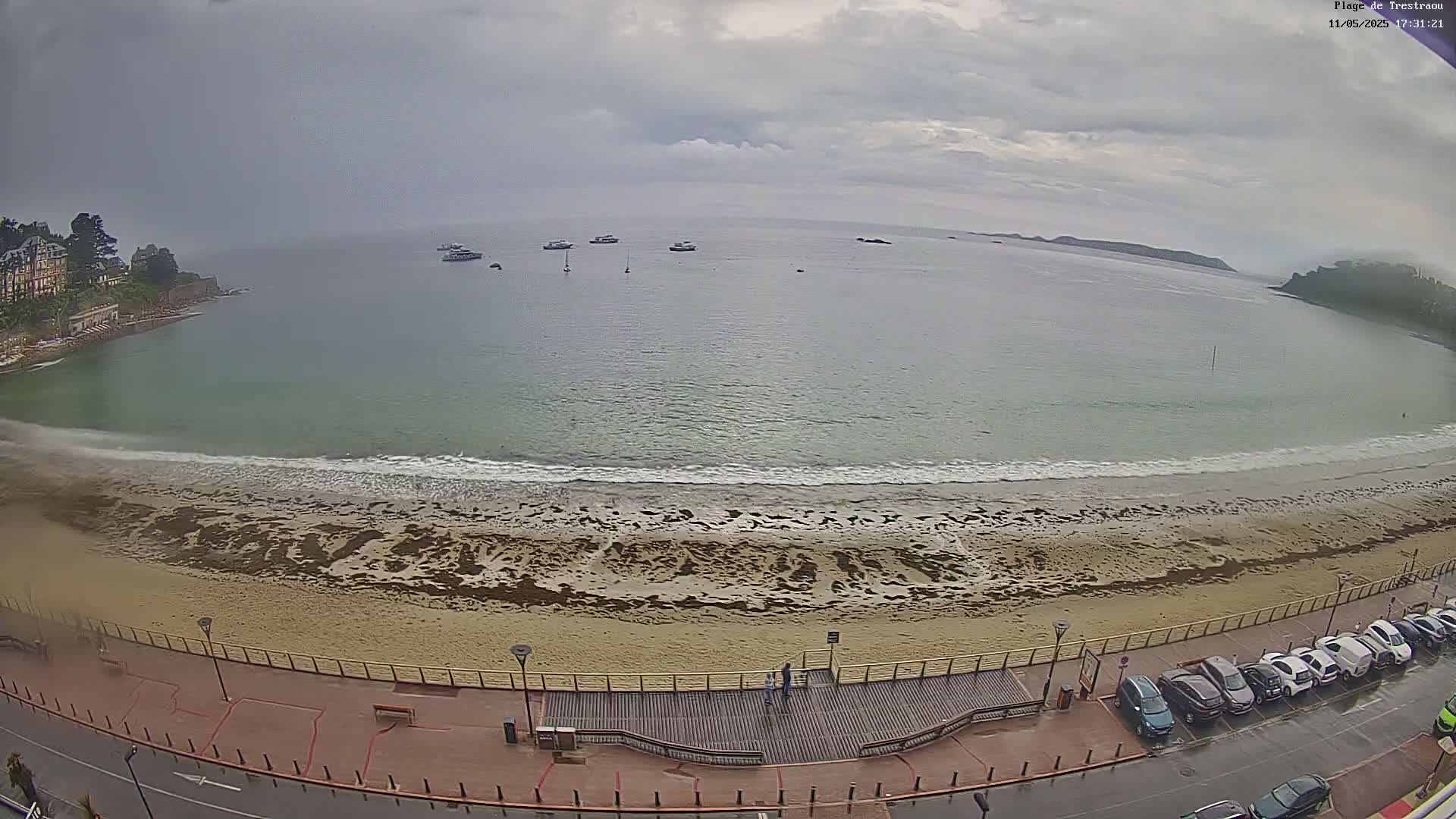 An overcast day shows a sandy beach with gentle waves lapping the shore, several boats in the calm bay, and a parking lot filled with cars next to a boardwalk.