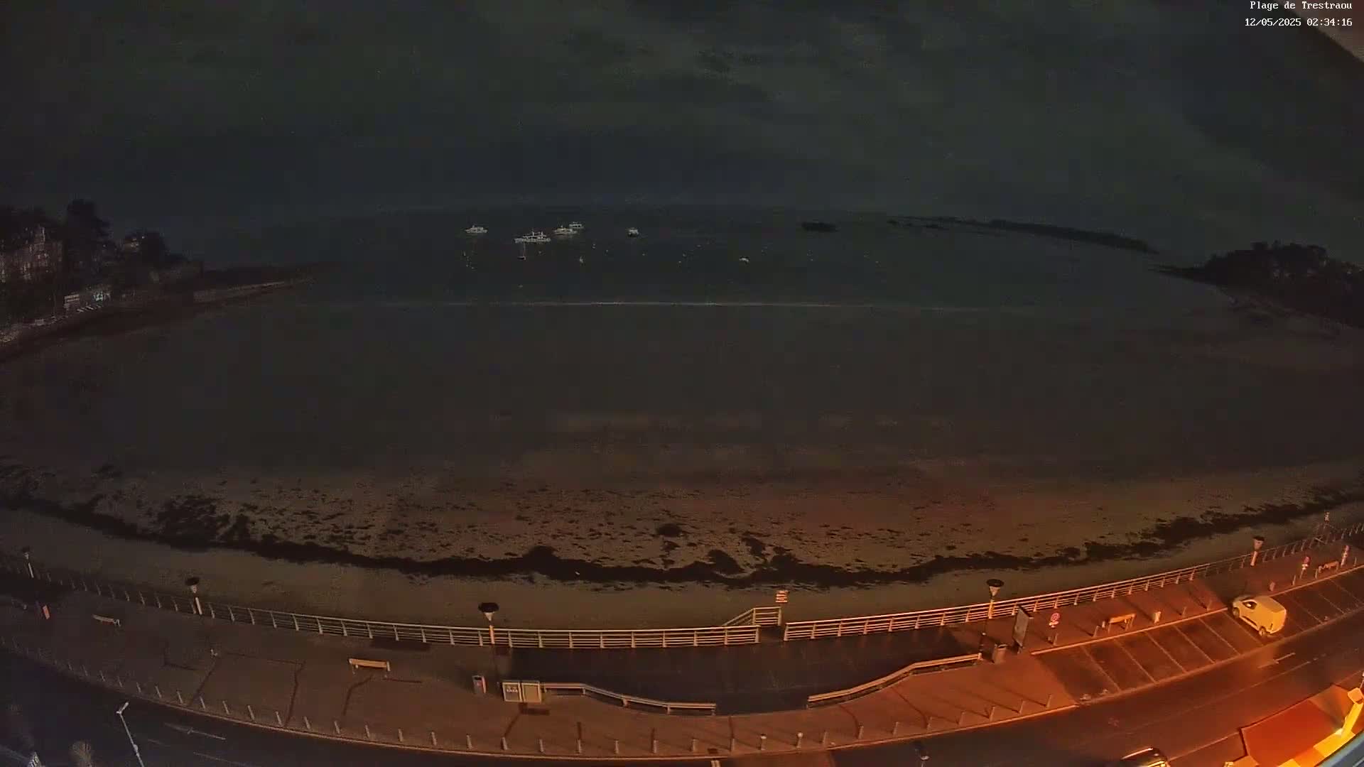 A dark, nighttime view of a beach with a few boats on the water and a mostly empty parking lot.