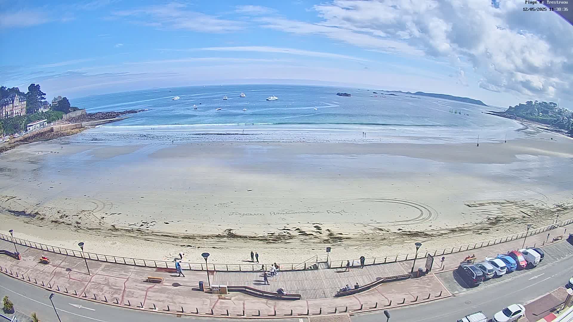 A sandy beach with a boardwalk and parked cars stretches along a calm ocean under a mostly sunny sky.