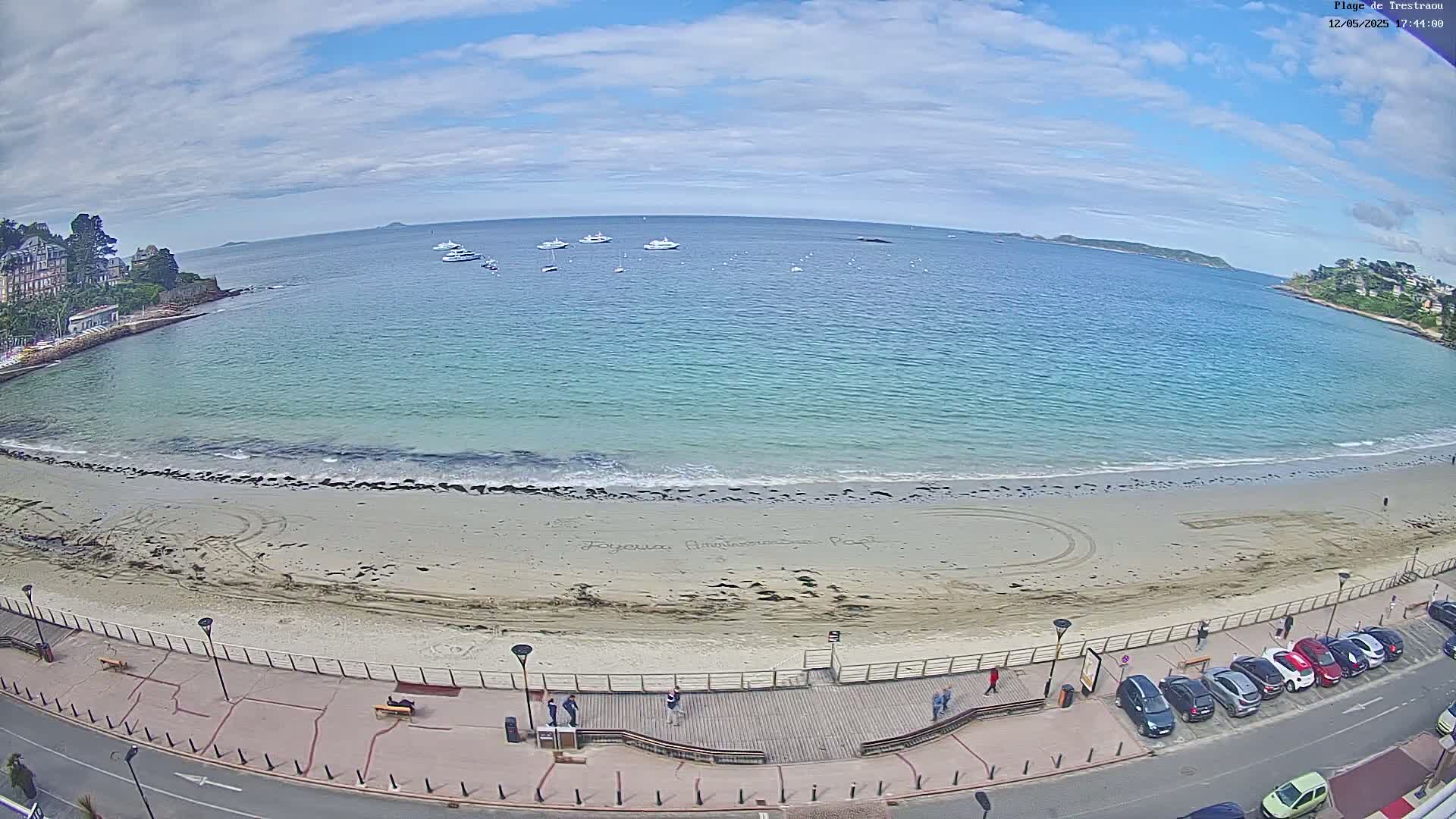 A sandy beach with a calm, light-blue ocean under a partly cloudy sky has several boats anchored in the distance, with a boardwalk and parked cars visible along the shore.