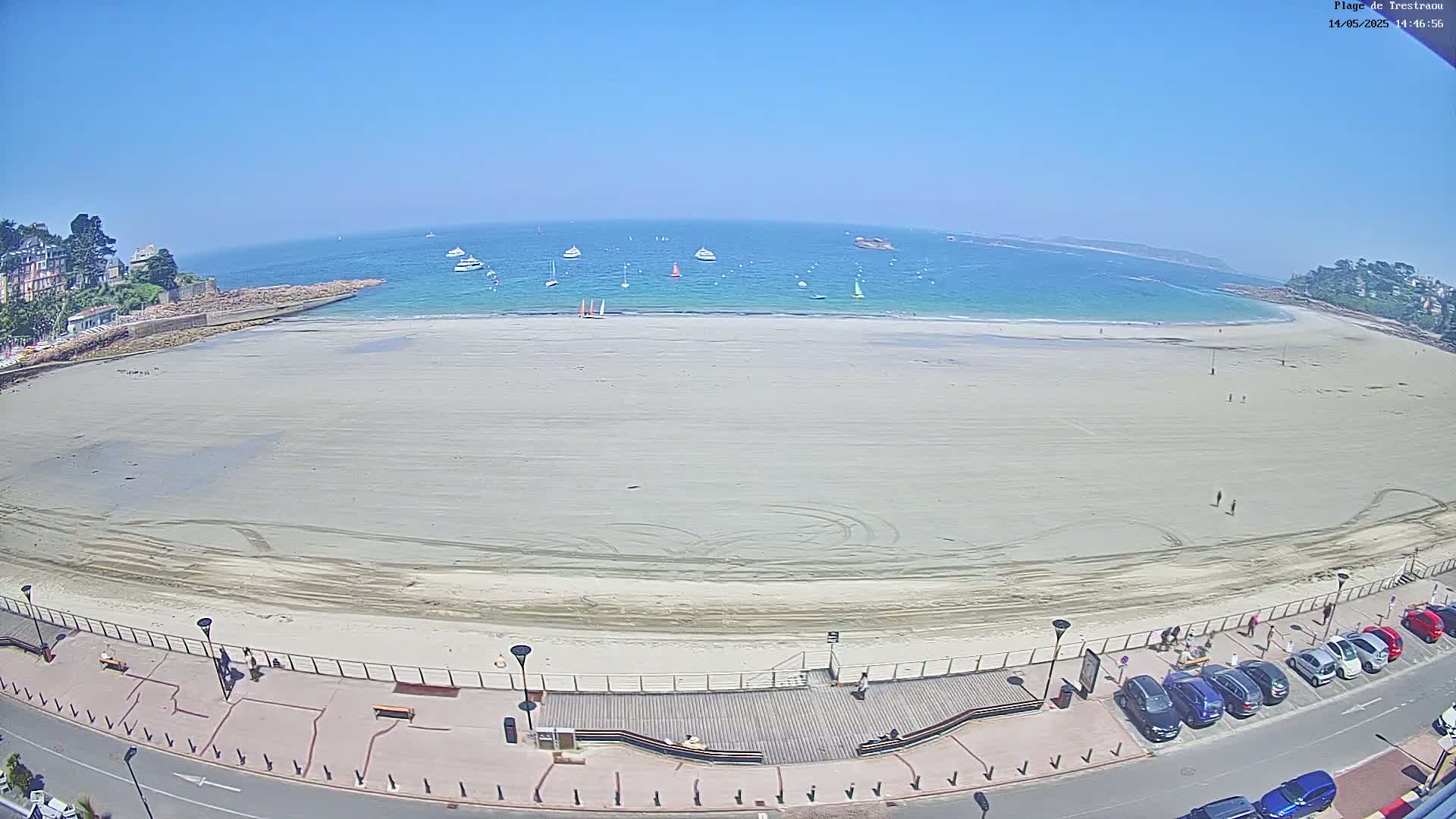 A wide sandy beach stretches along a calm, turquoise ocean under a clear blue sky, with several boats visible on the water and cars parked along the beachfront boardwalk.