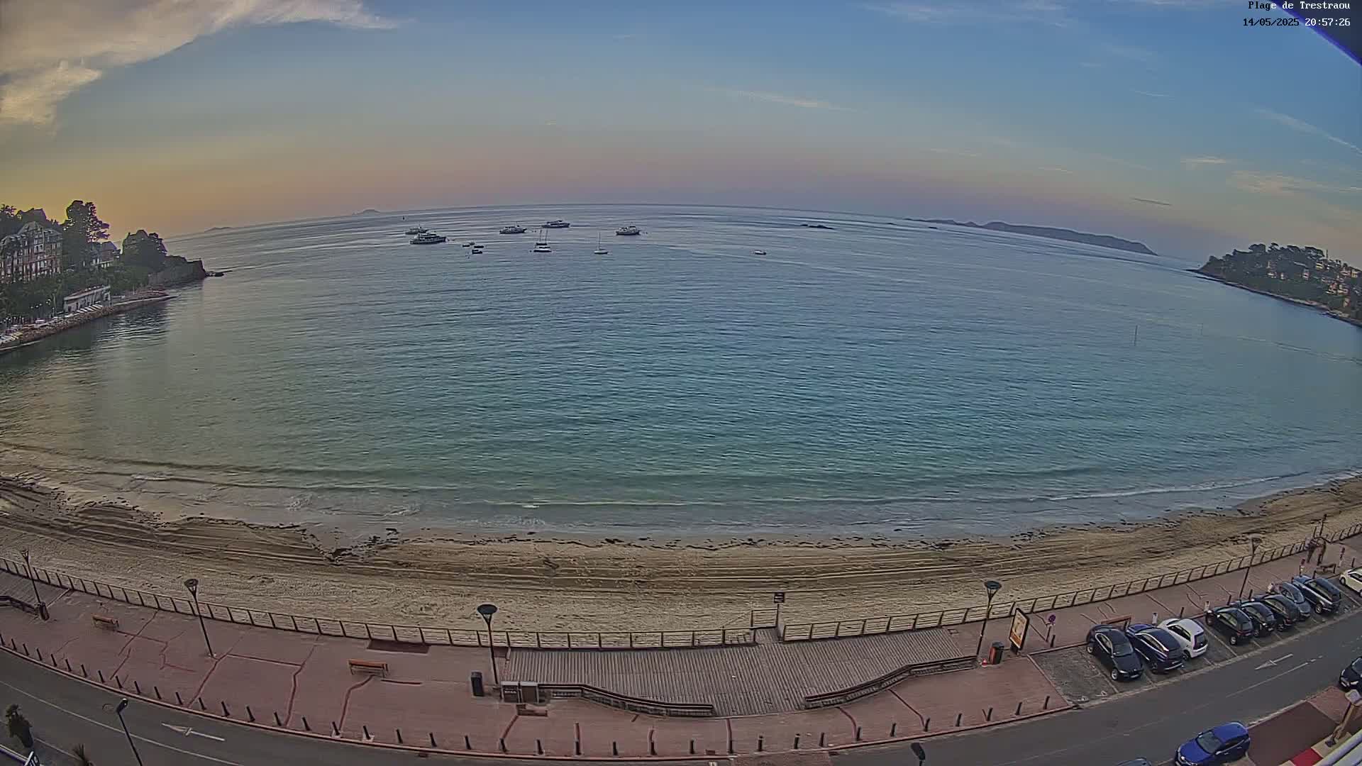 A sandy beach with calm, clear water and several boats in a bay at sunset, bordered by buildings and a parking lot.