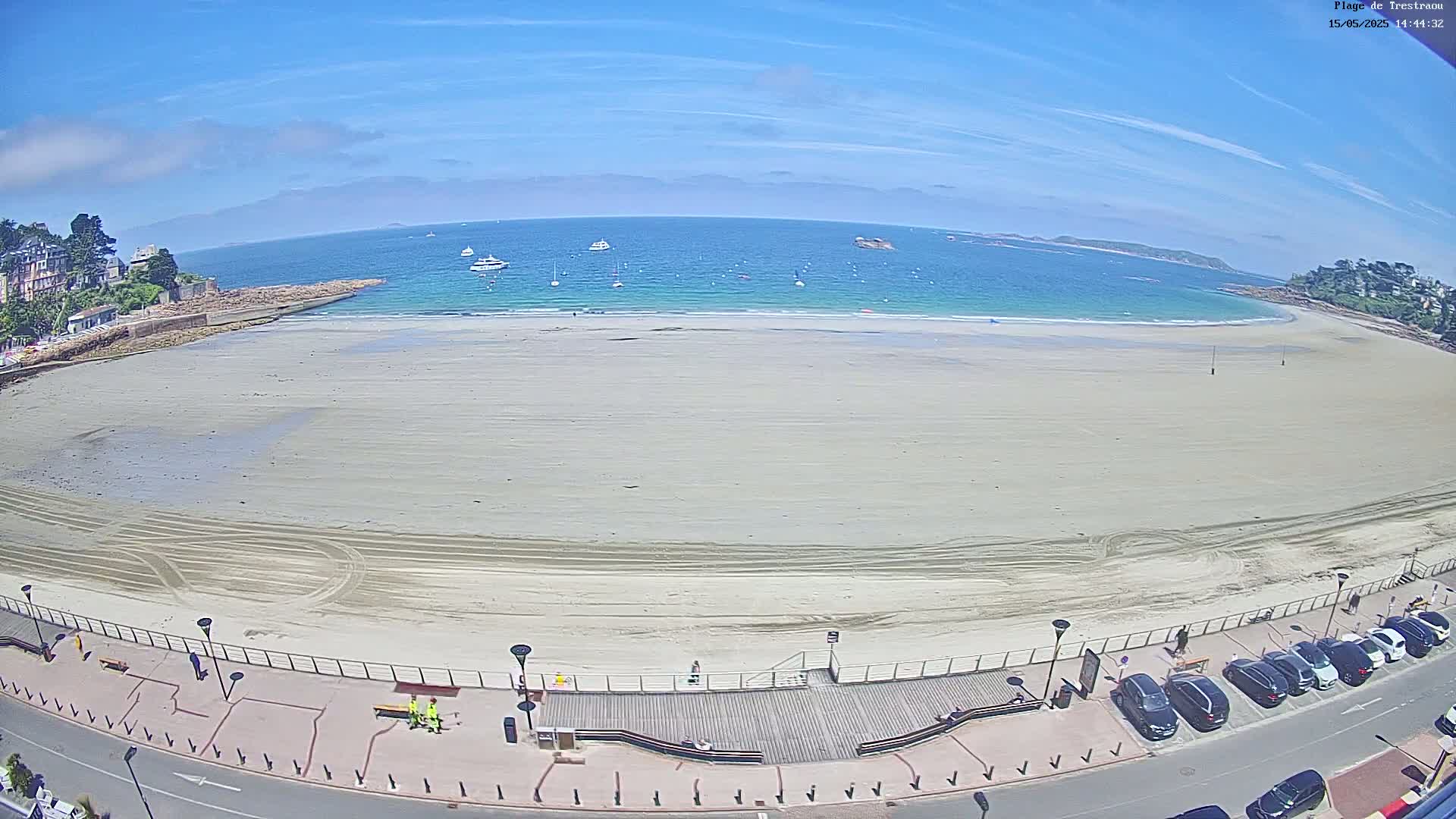 A mostly empty sandy beach stretches along a calm, turquoise ocean under a bright, sunny sky, with several boats visible in the distance and cars parked along the road bordering the beach.