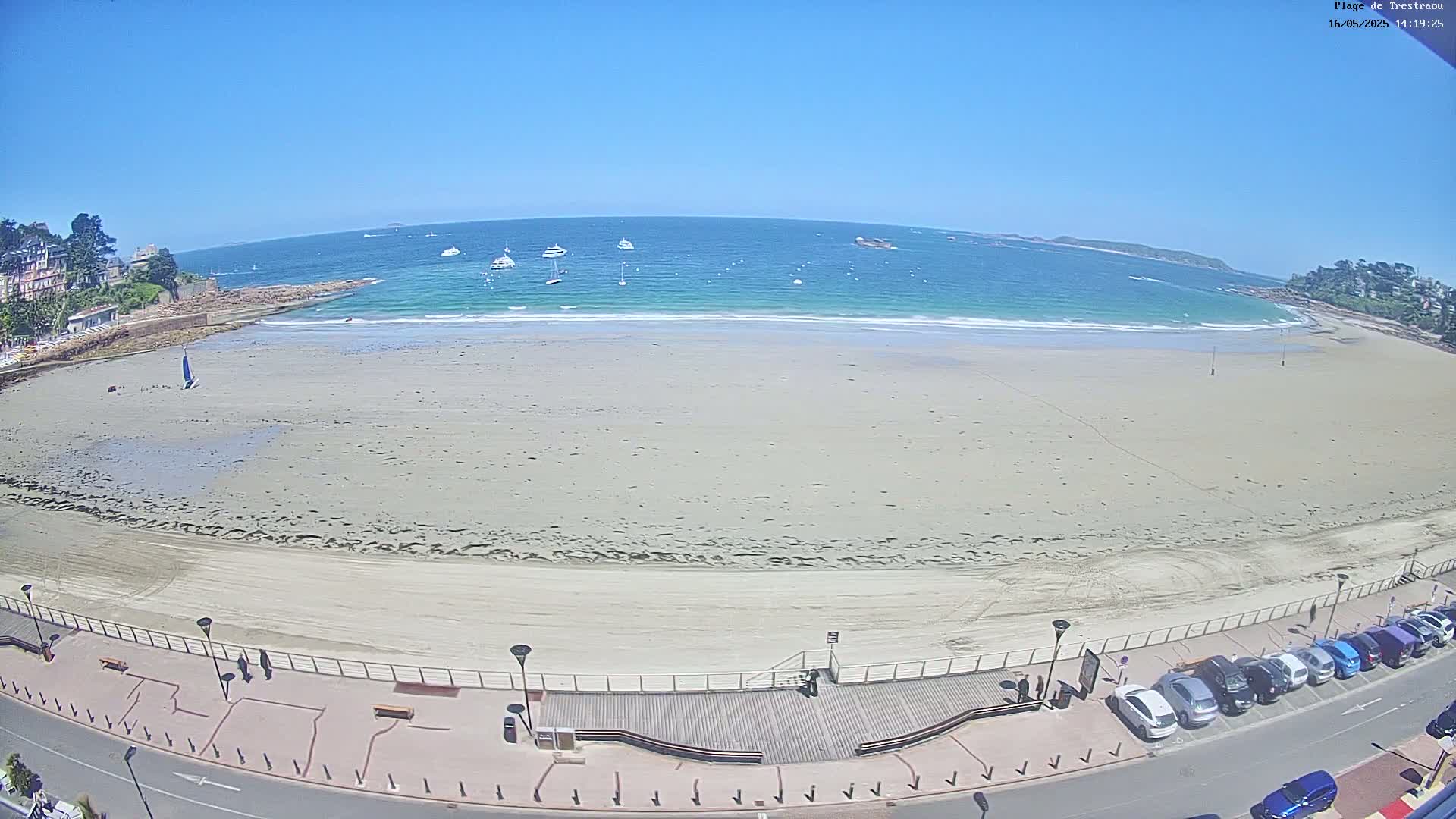 A wide sandy beach stretches out to a calm, turquoise ocean under a clear blue sky, with several boats visible on the water and cars parked along a paved road bordering the beach.