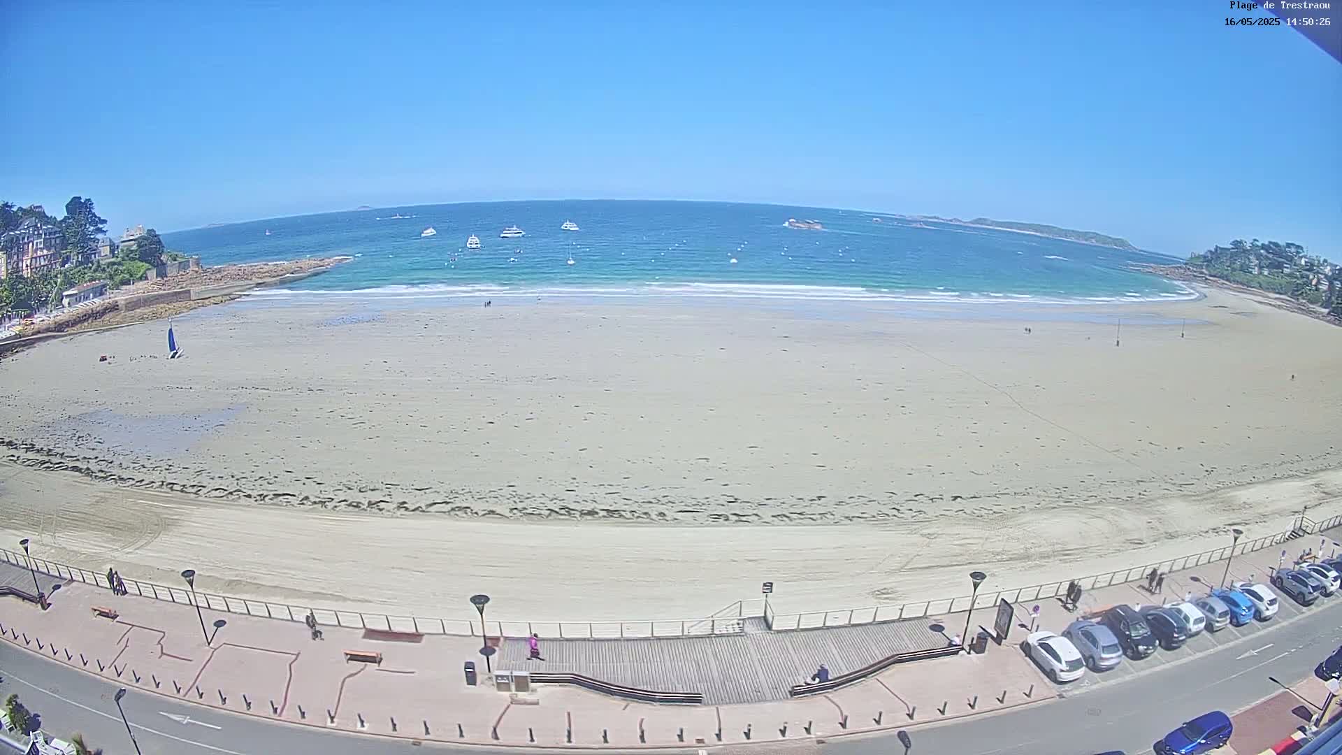 A wide sandy beach with a few people and several boats in the distance on a sunny day.