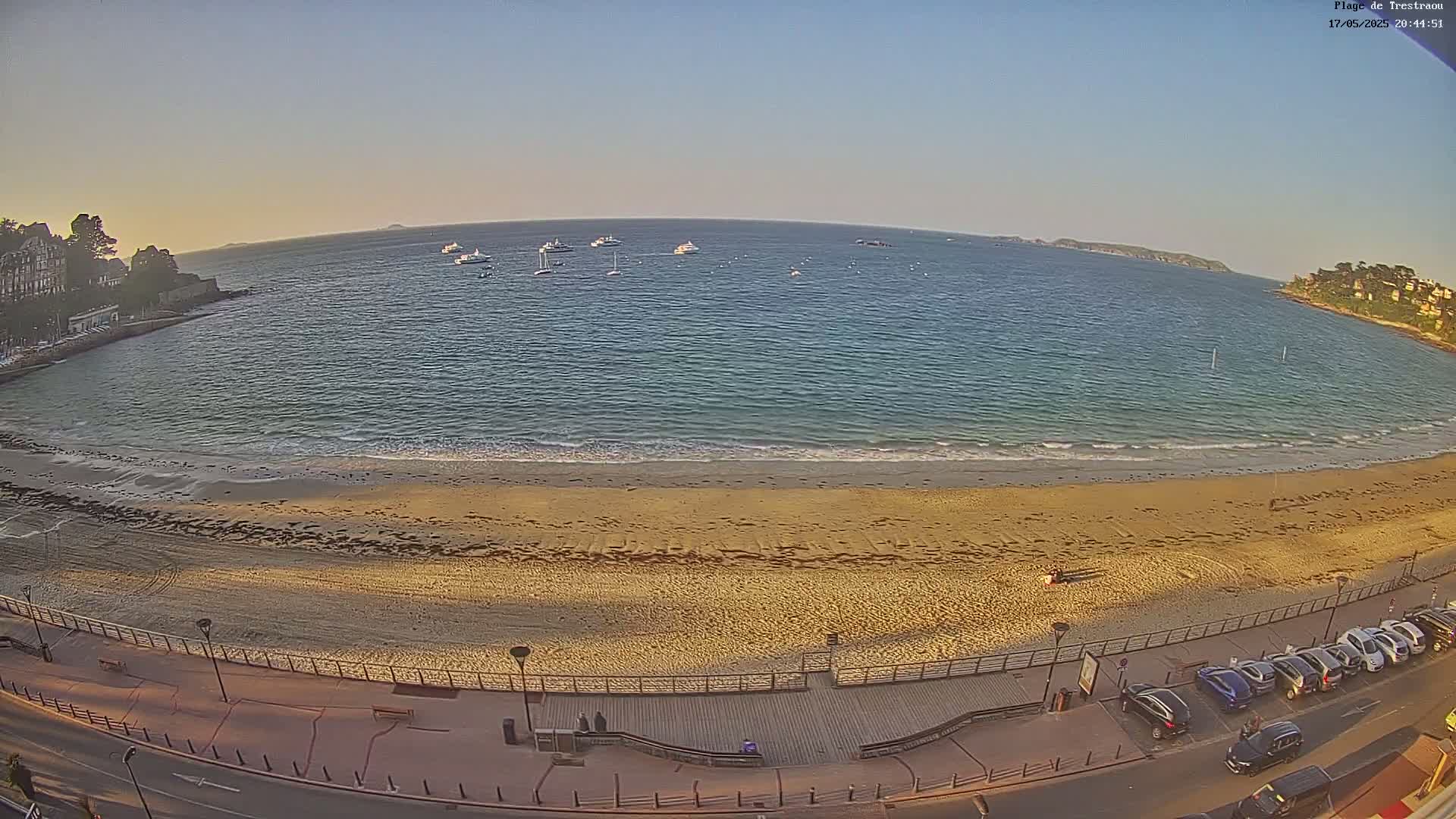 A sandy beach with calm ocean waves and several boats in the distance under a clear, bright sky.