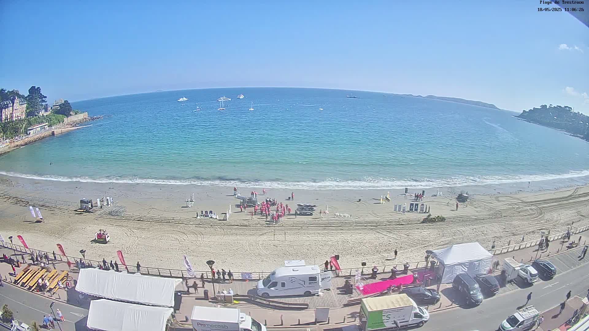 A sandy beach with people and vehicles along the shoreline, under a clear blue sky, overlooks a calm, teal ocean with several boats visible in the distance.
