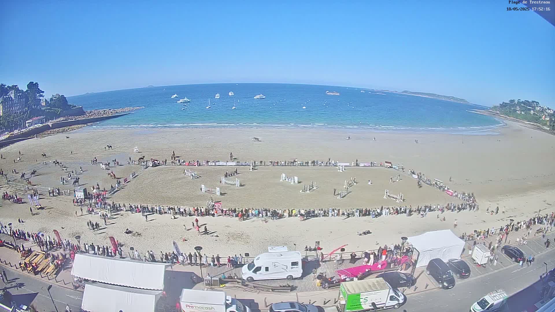 A sunny day on a sandy beach shows an equestrian competition in progress, with numerous horses and riders, spectators lining the course, and boats visible in the calm ocean beyond.