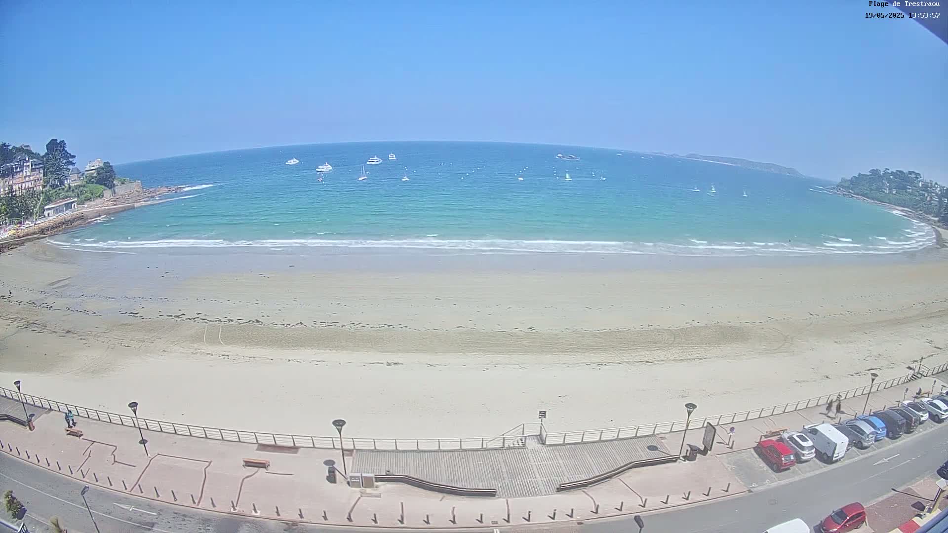 A sandy beach with calm, light-blue water under a clear blue sky, boats are visible in the distance, and cars are parked along the road next to the beach.