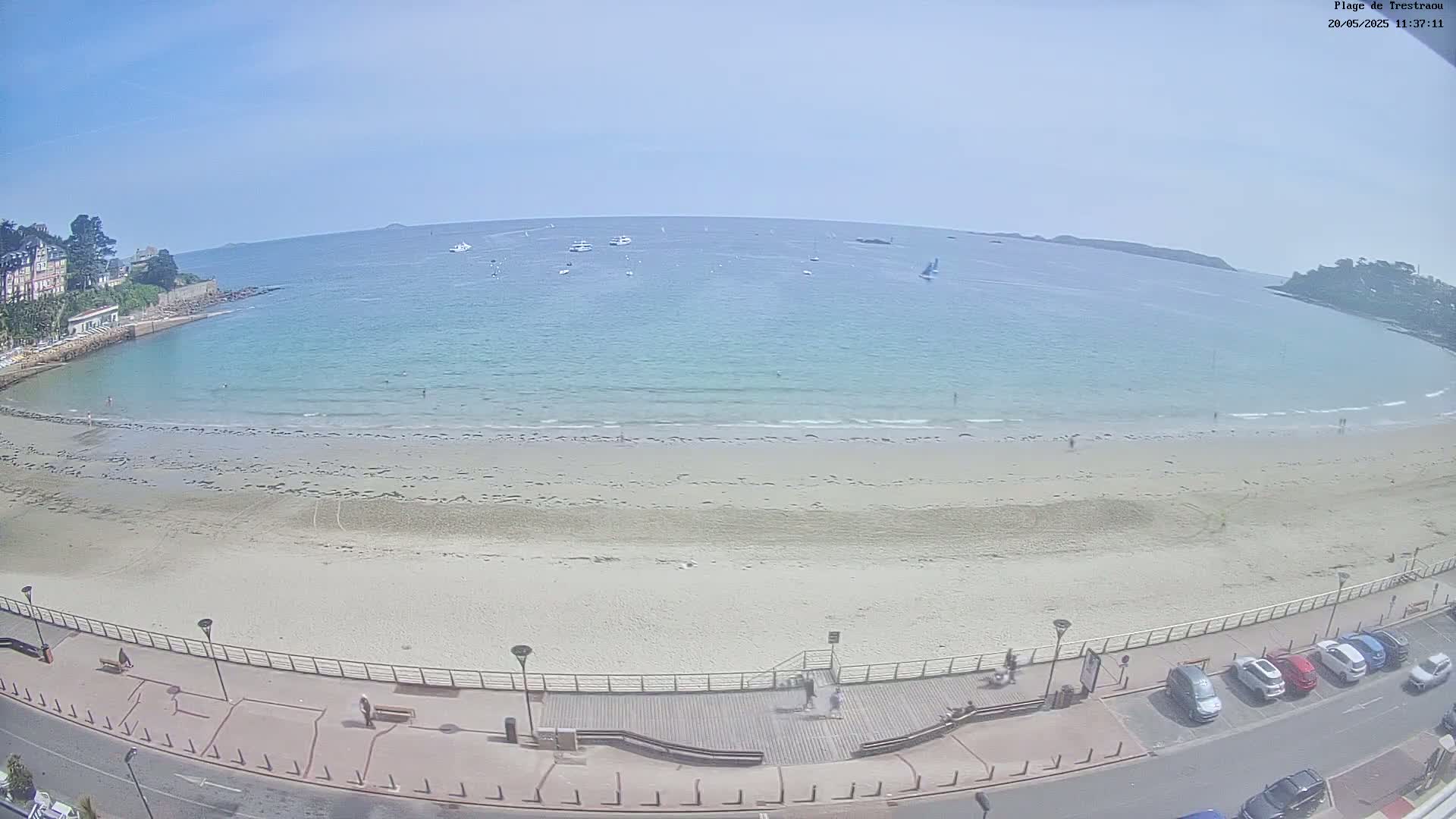 A sandy beach with calm, light-blue water stretches out to a horizon under a clear blue sky, featuring several boats in the distance and cars parked along a road next to the beach.