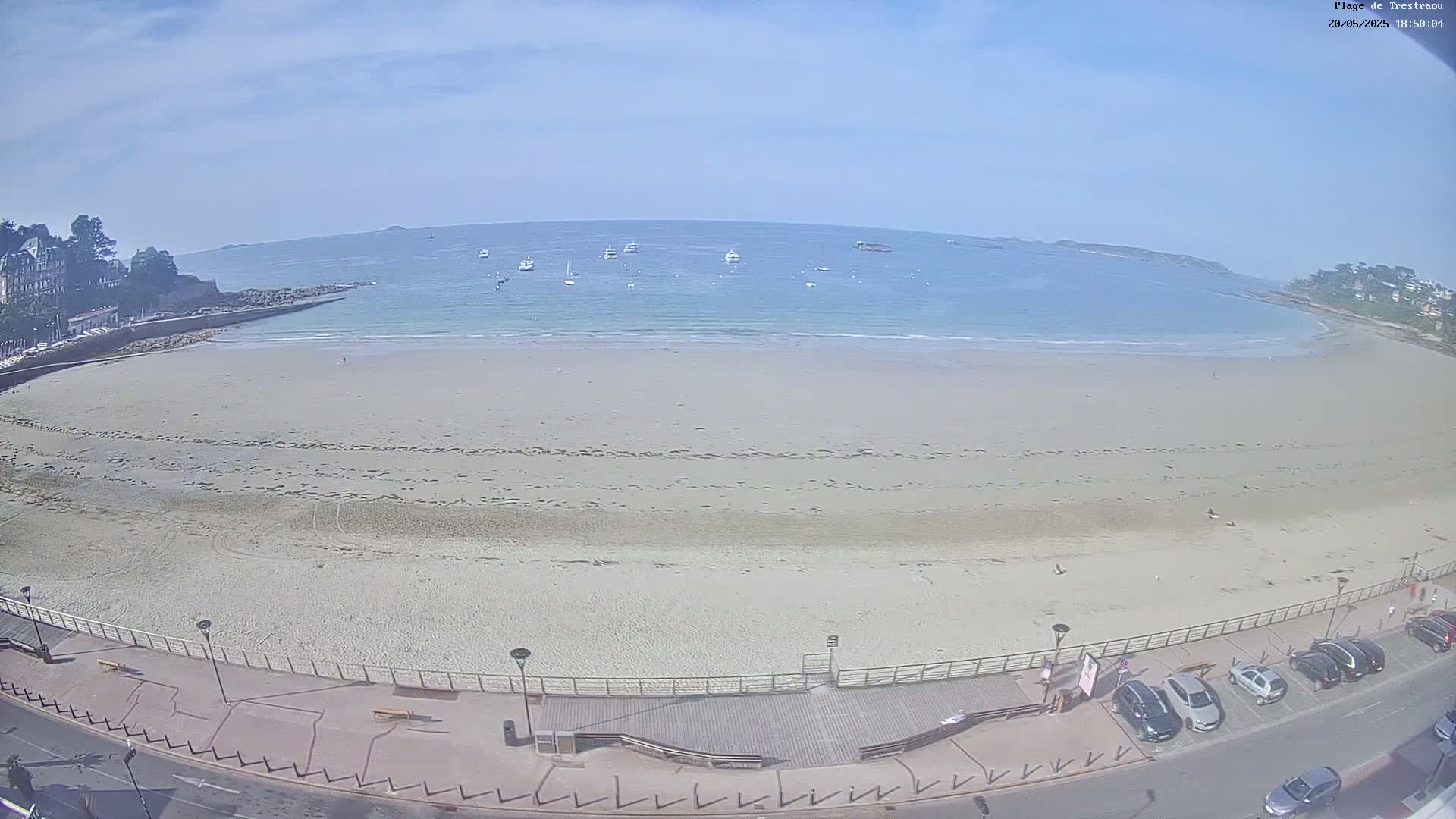 A wide sandy beach with a calm ocean and several boats in the distance under a mostly clear sky is seen from an elevated vantage point.
