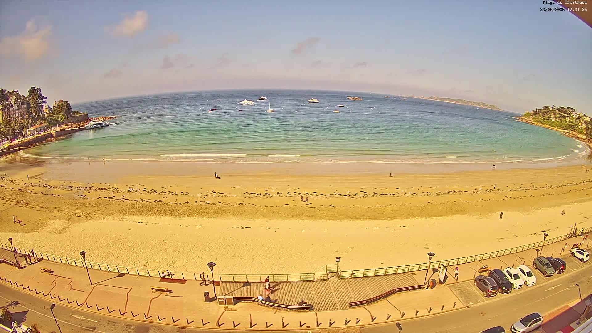 A sandy beach with calm, teal water curves around a seaside town under a mostly sunny sky.