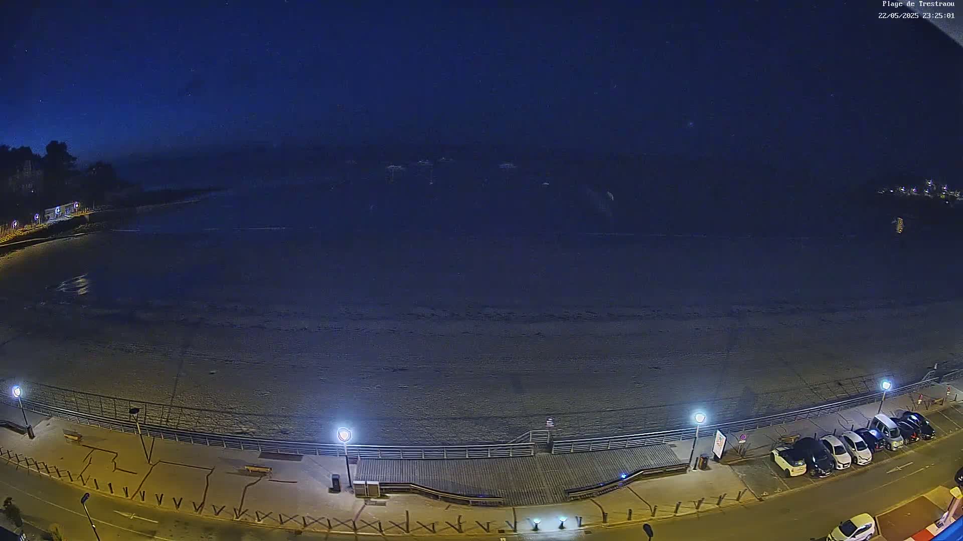 A nighttime, overhead view shows a mostly empty beach with a boardwalk, street lights, and several cars parked along a road.