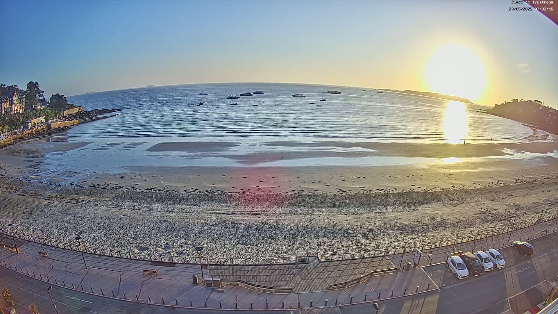 A sandy beach with calm waves and several boats in the distance is bathed in the golden light of a rising sun.