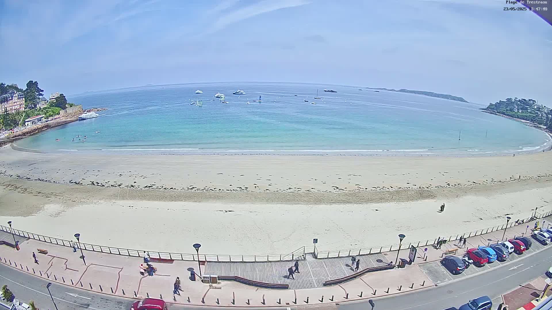A wide, sandy beach with turquoise water and several boats visible in the distance under a clear, sunny sky is bordered by a road with parked cars and a walkway with people walking.