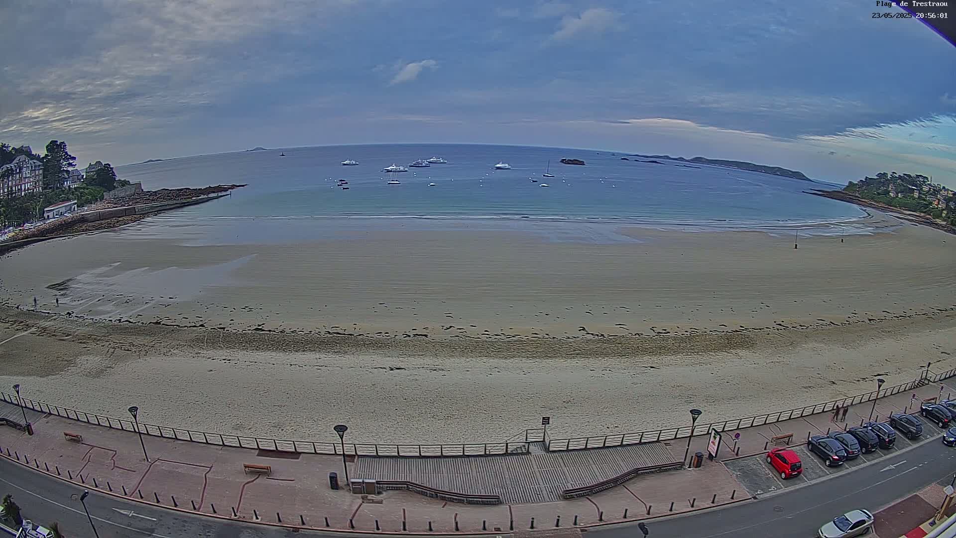 An expansive sandy beach, bordered by a roadway and buildings, stretches along a calm bay with several boats anchored in the water under a cloudy sky.