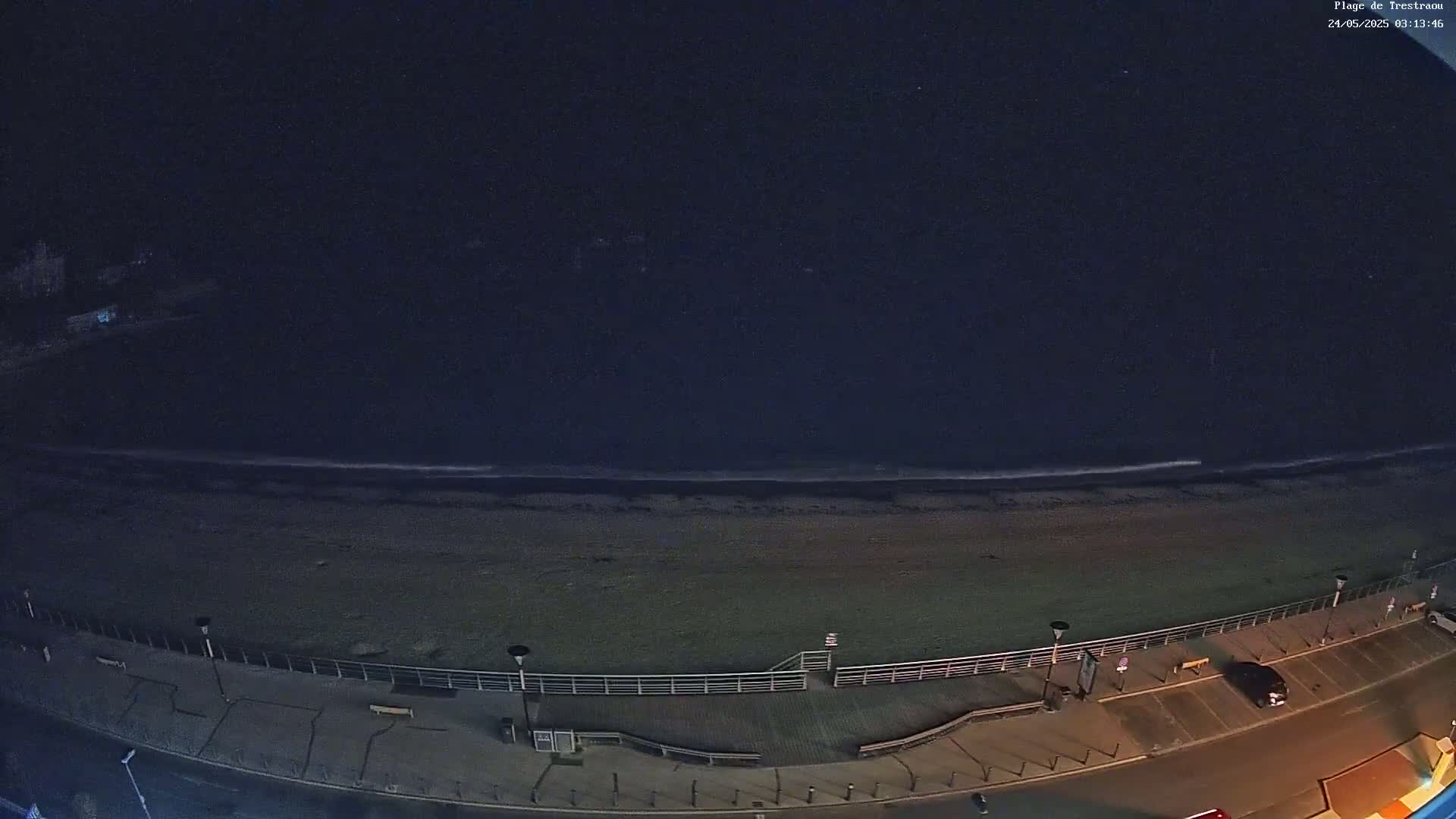 A dark, nighttime view of a beach with a calm ocean, a boardwalk, and a few parked cars.