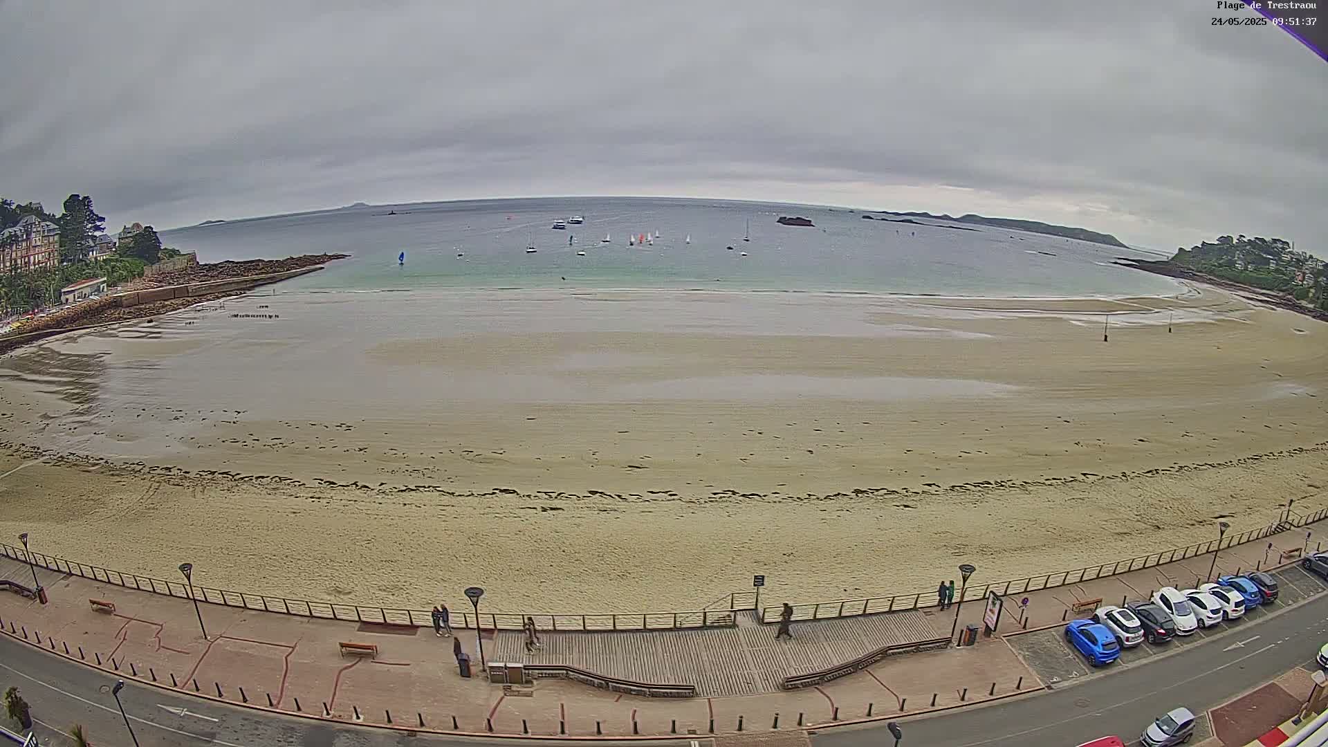 A wide shot of a sandy beach with several sailboats in the calm water, under an overcast sky.