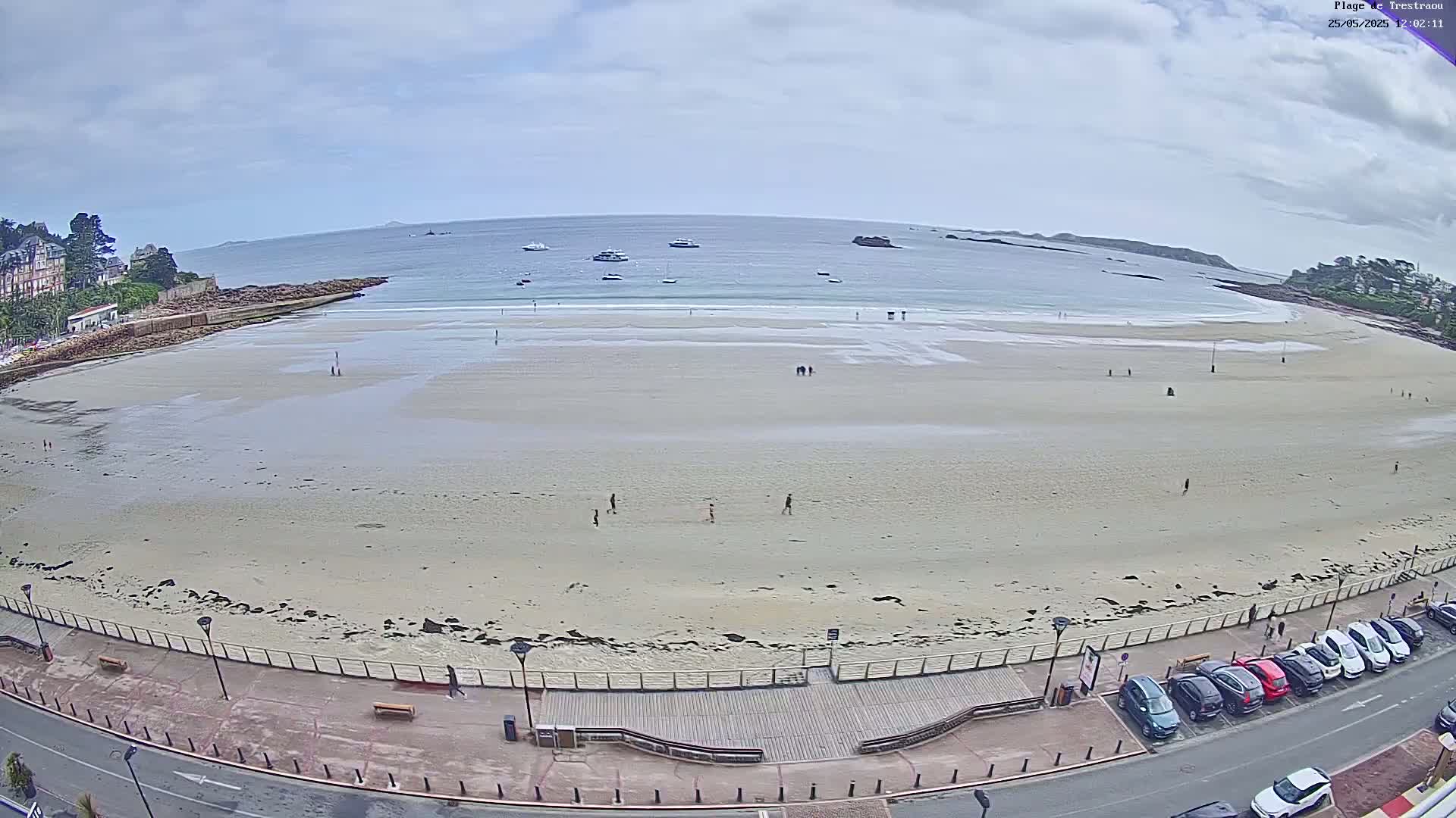 A mostly empty sandy beach stretches along a calm ocean under a cloudy sky, with several people scattered across the sand and a row of parked cars visible along the adjacent road.