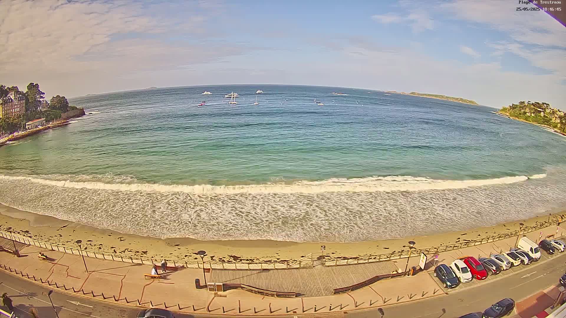 A sandy beach with gentle waves laps the shore under a partly cloudy sky, with several boats visible in the calm water beyond.