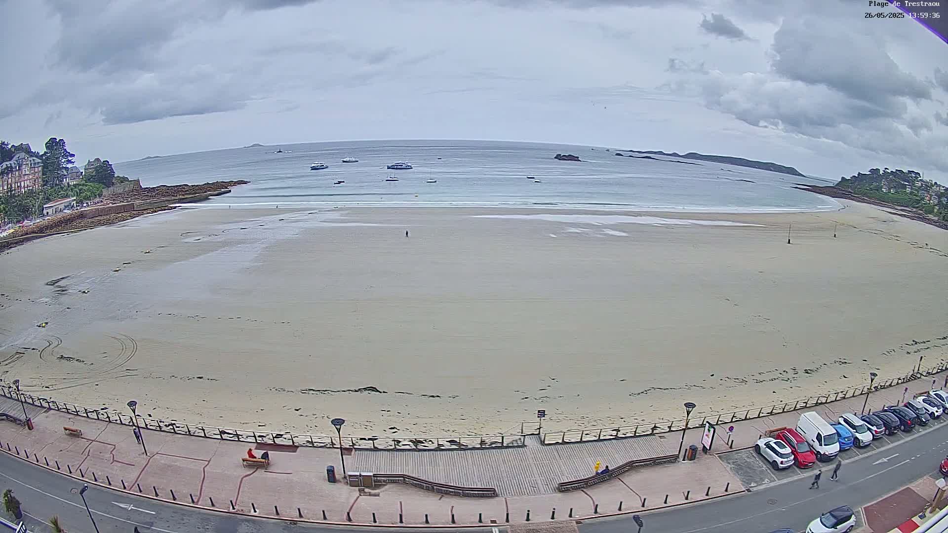A wide sandy beach stretches to a calm sea under a cloudy sky, with several boats visible in the distance and cars parked along a roadway bordering the beach.
