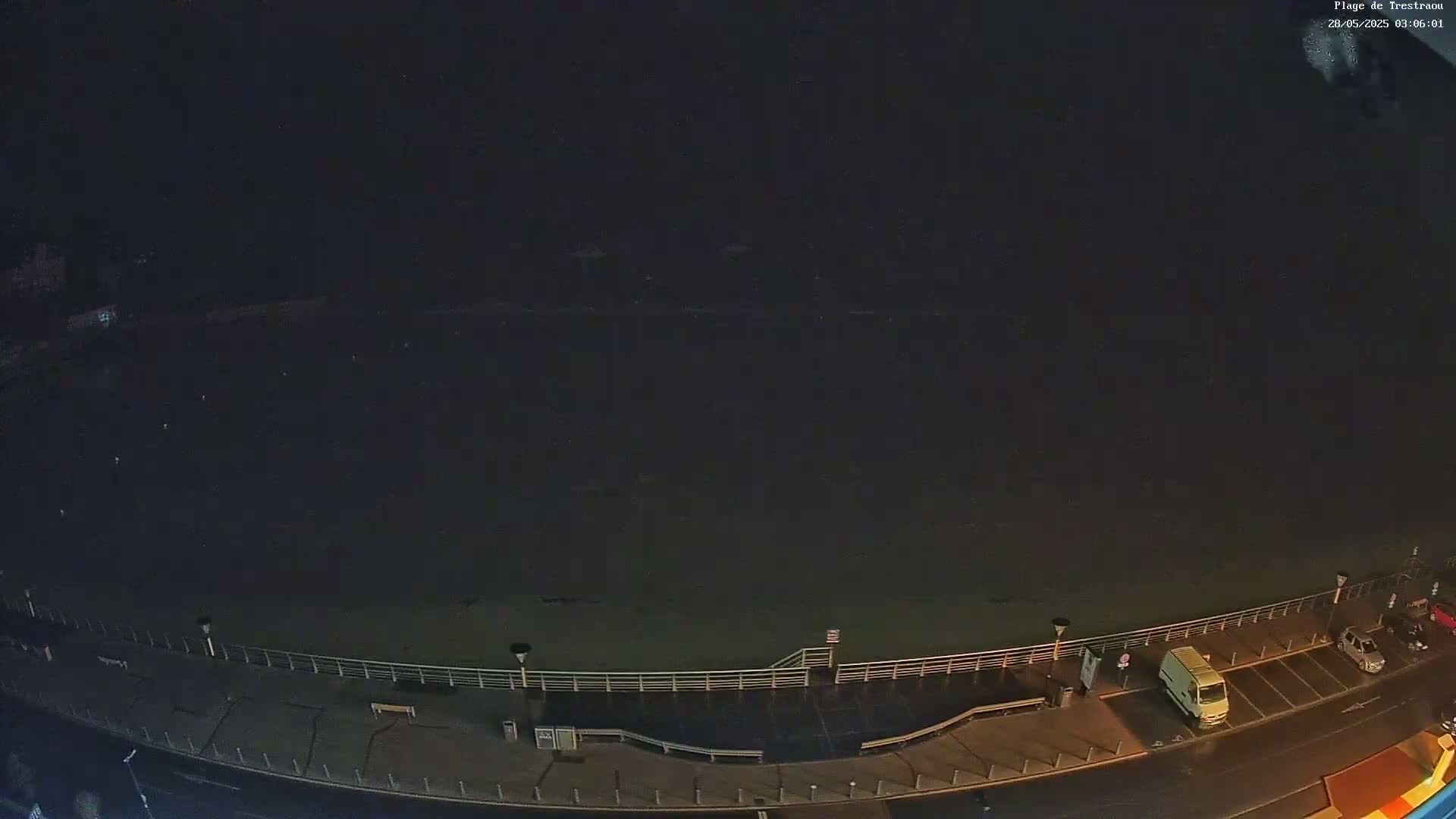A dark, nighttime view shows a mostly empty beach, a paved walkway with railings, and a few vehicles parked in a lot next to the road.