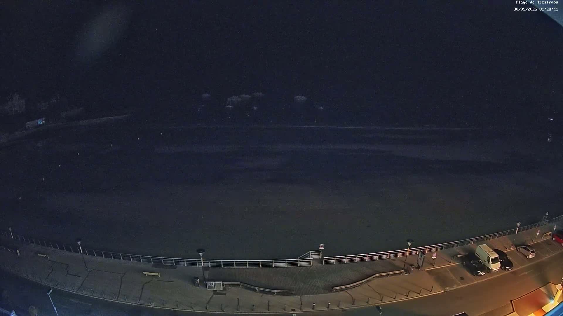 A dark, nighttime view of a mostly empty beach with a boardwalk, a few parked cars, and some distant lights.