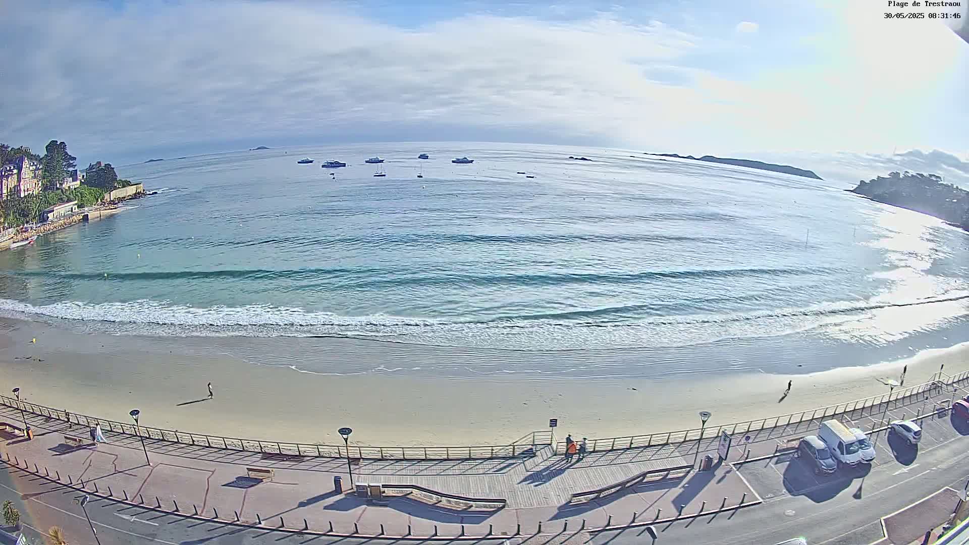 A sandy beach with gentle waves laps the shore under a partly cloudy sky, several boats are visible in the calm water beyond.